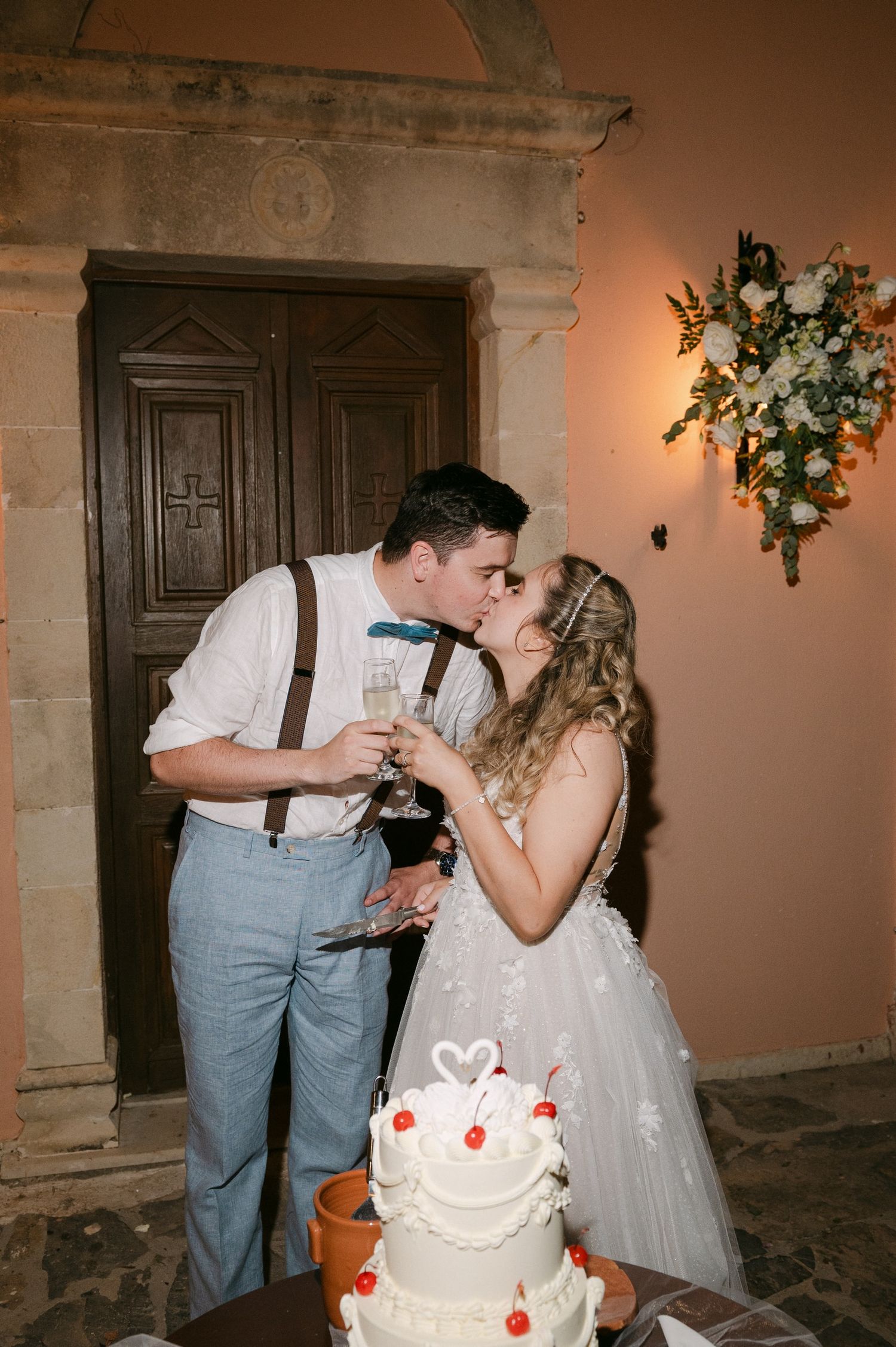A couple shares a romantic kiss while cutting their white wedding cake adorned with red hearts.