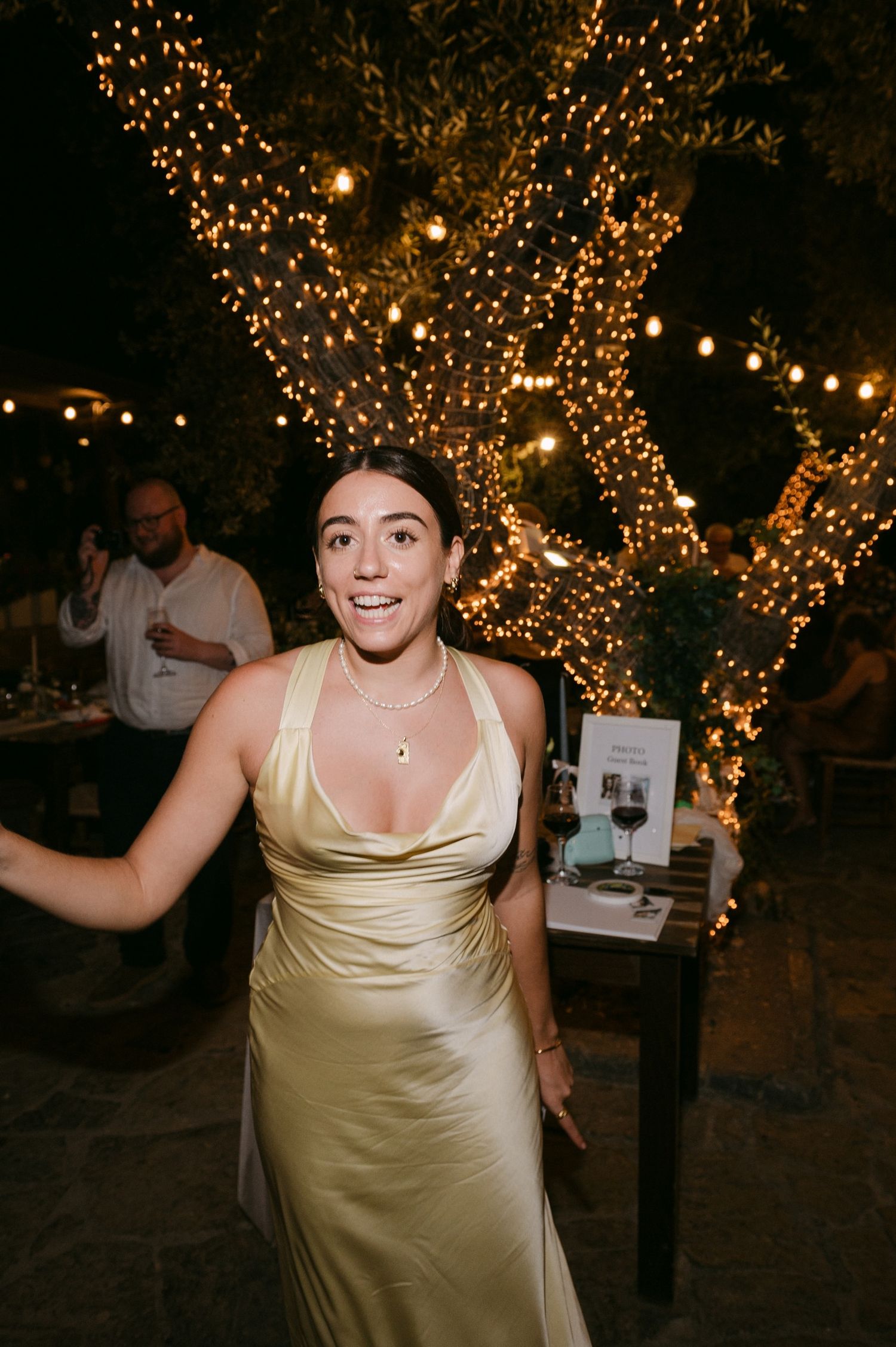 A person in a champagne-colored satin dress dances joyfully at a nighttime celebration under twinkling string lights.