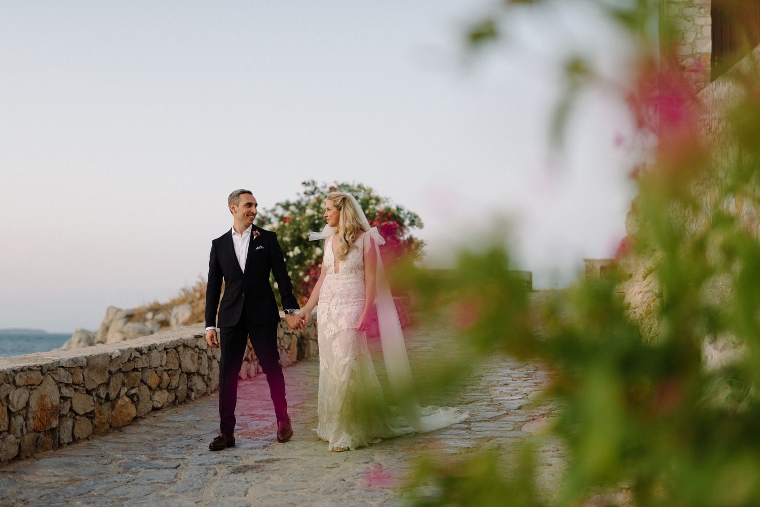 Wedding couple walks hand in hand on stone path with pink flowers and Mediterranean scenery.