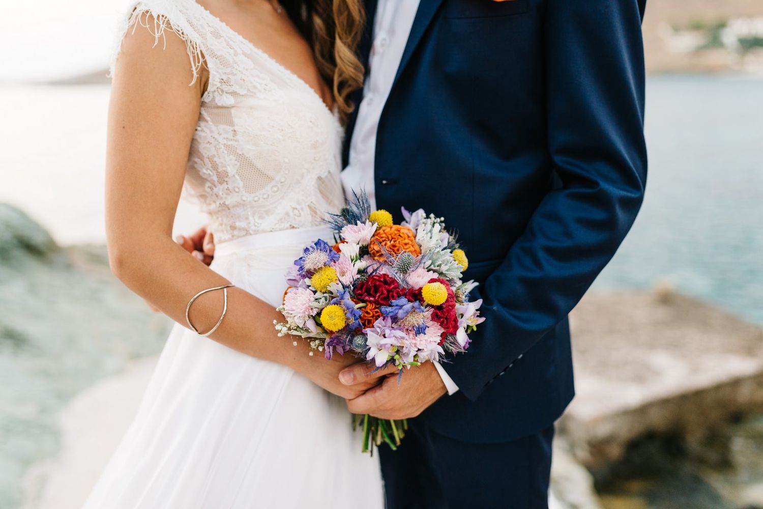 Close-up of couple holding colorful wedding bouquet against coastal background.