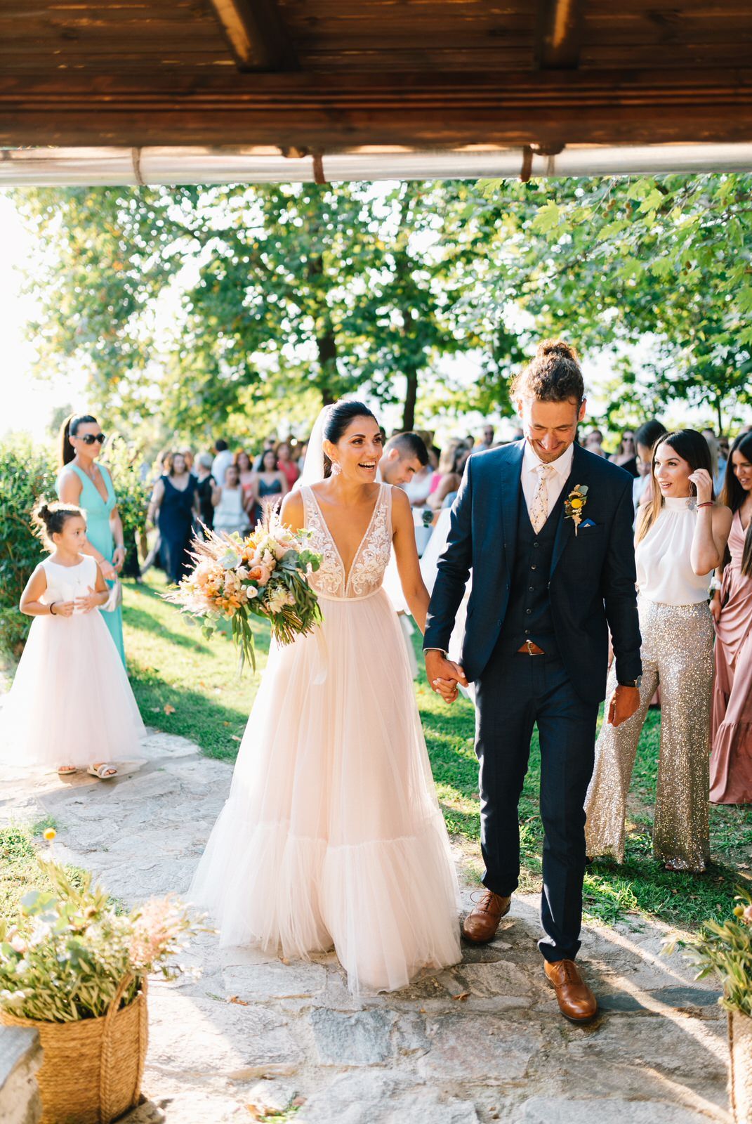 Newlyweds walk together along a covered pathway surrounded by wedding guests.