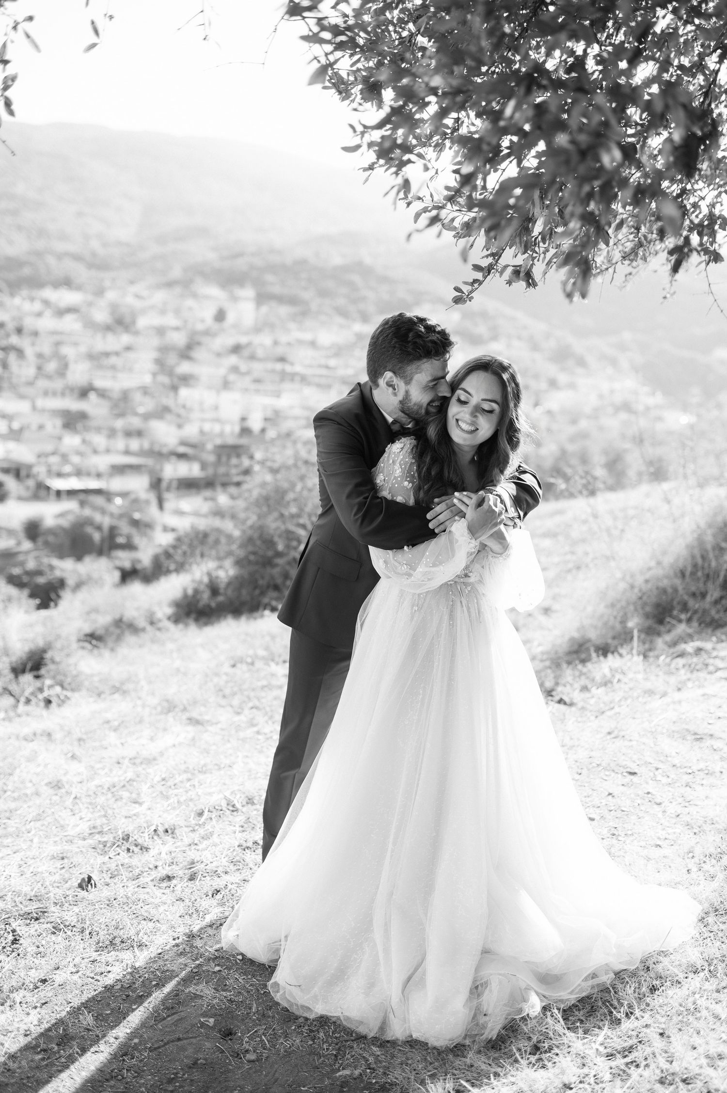 A romantic black and white wedding photo captured on a hillside overlooking a distant town.