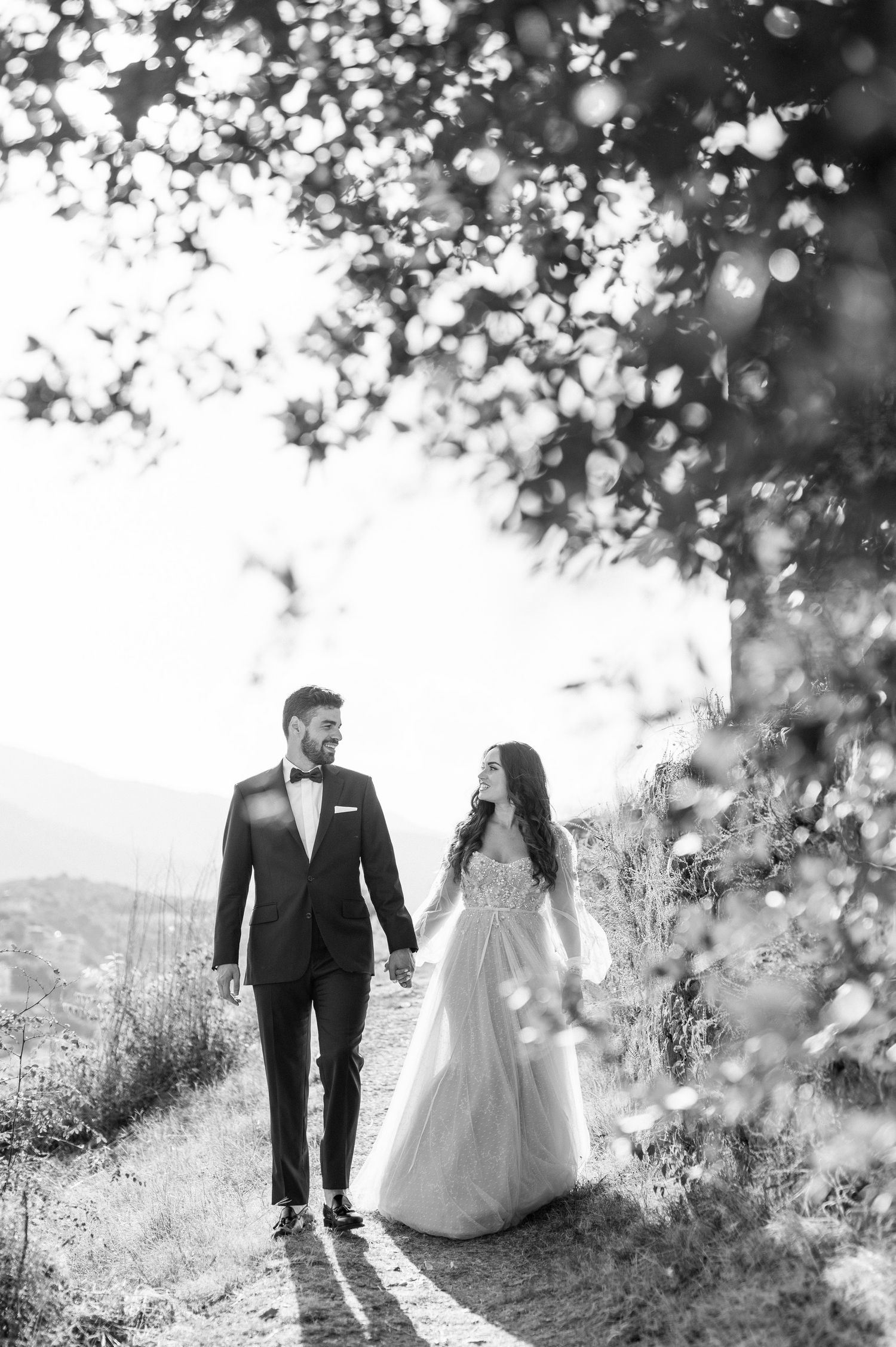 A black and white wedding photo shows a couple walking hand-in-hand along a sunlit mountain path.