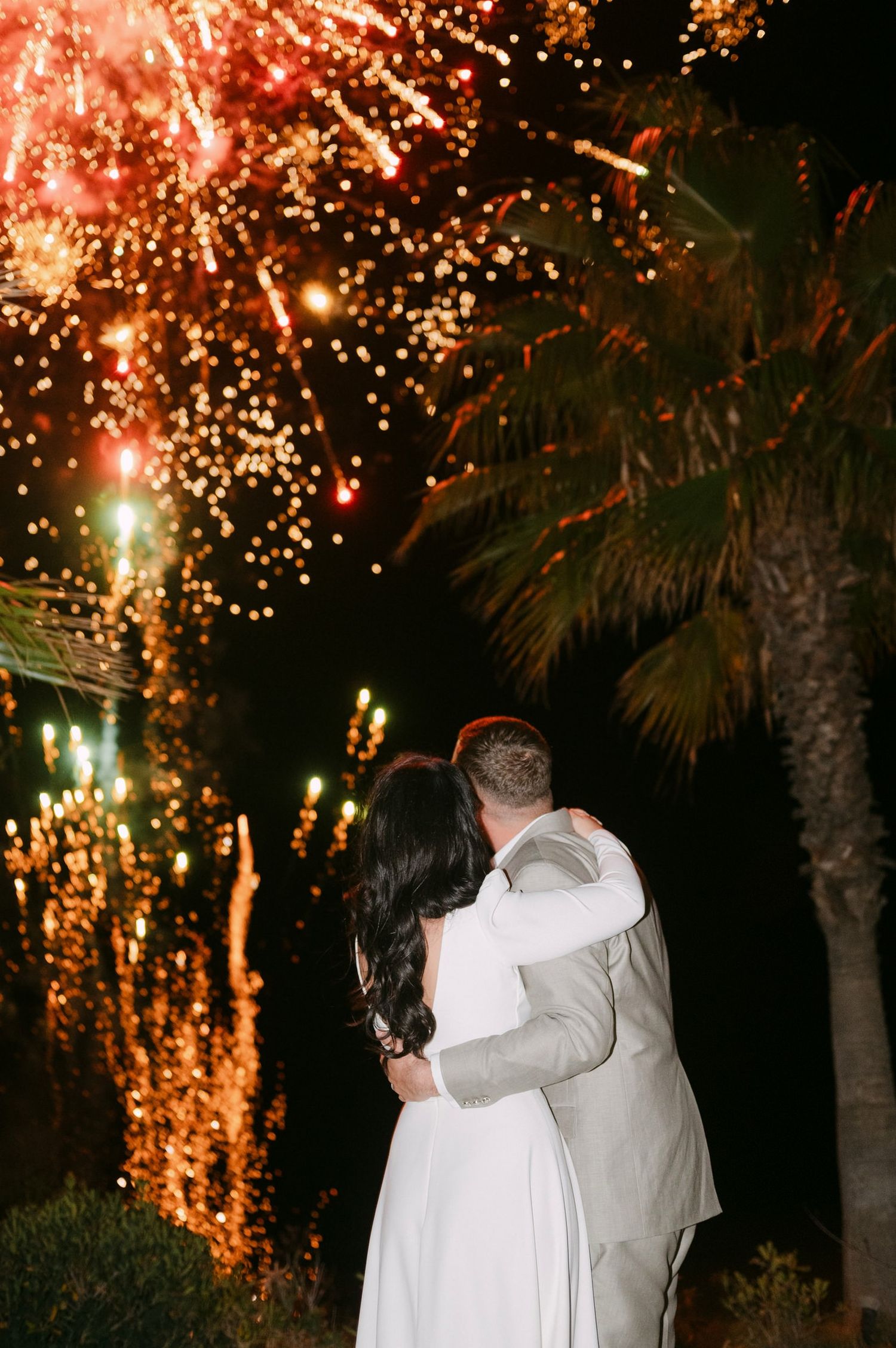 A couple embraces under fireworks at night with palm trees in the background creating a romantic wedding moment.