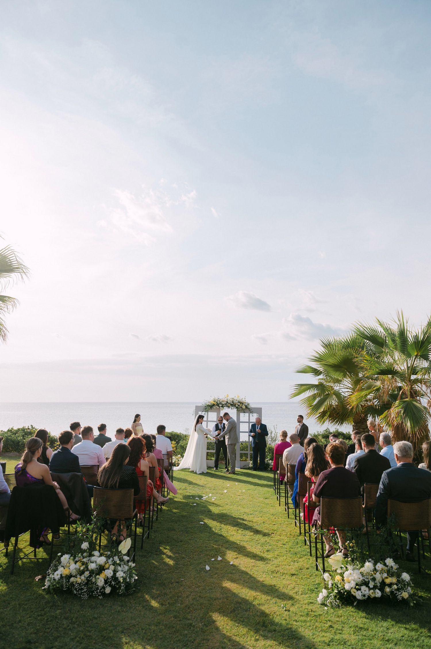 Guests seated at an outdoor wedding ceremony overlooking the ocean with palm trees and blue skies in the background.