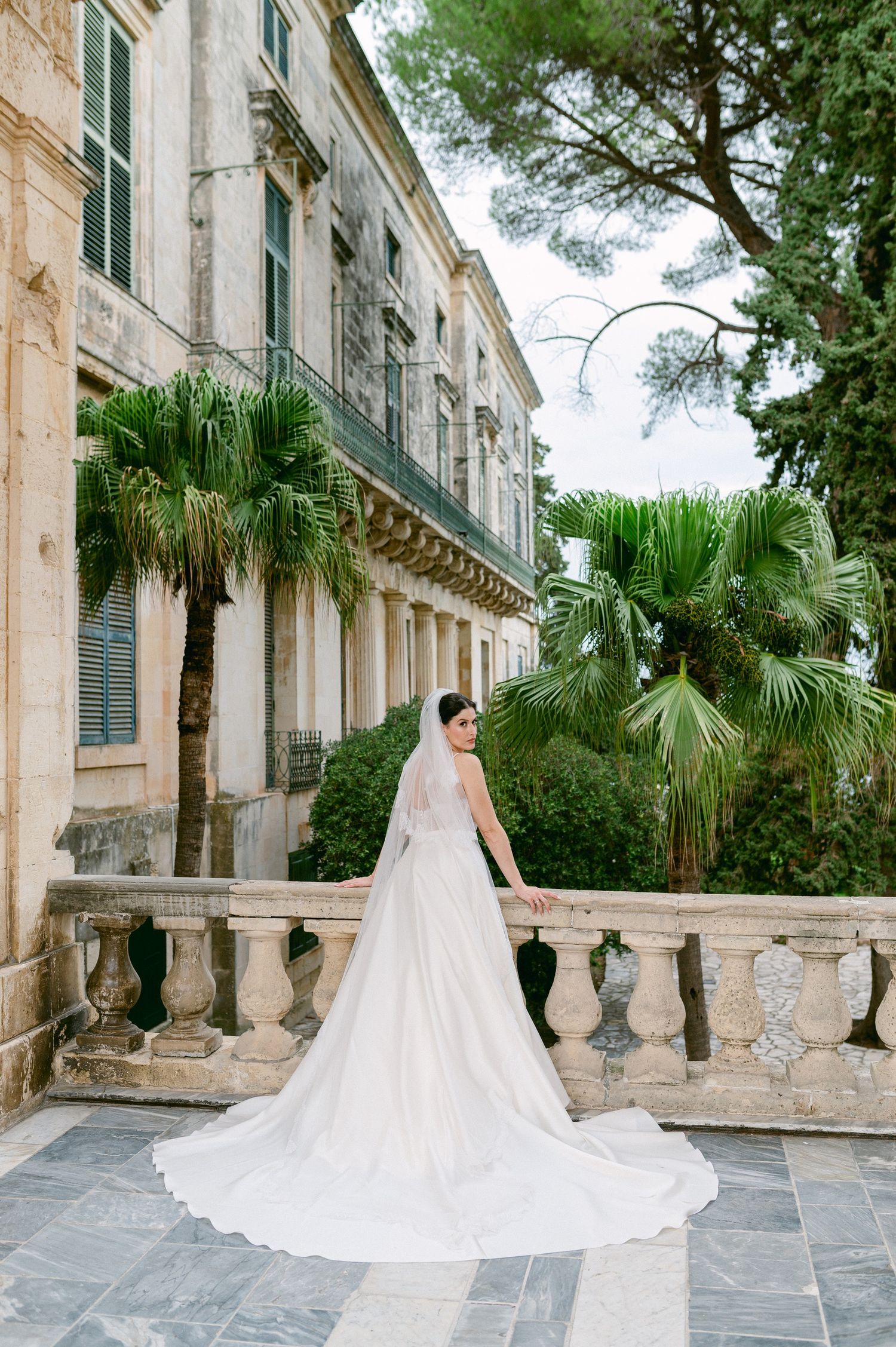A bride in an elegant white gown and flowing cathedral veil poses on a grand stone terrace with palm trees and columns in Corfu Greece