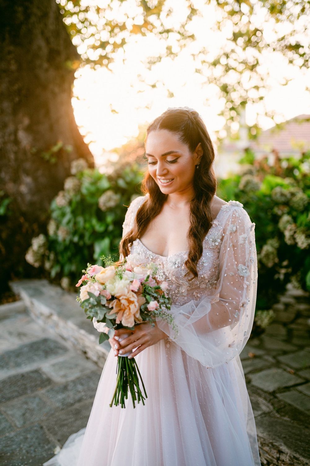 A bride in a delicate lace gown with billowing sleeves holds a soft peach and blush bouquet at golden hour.