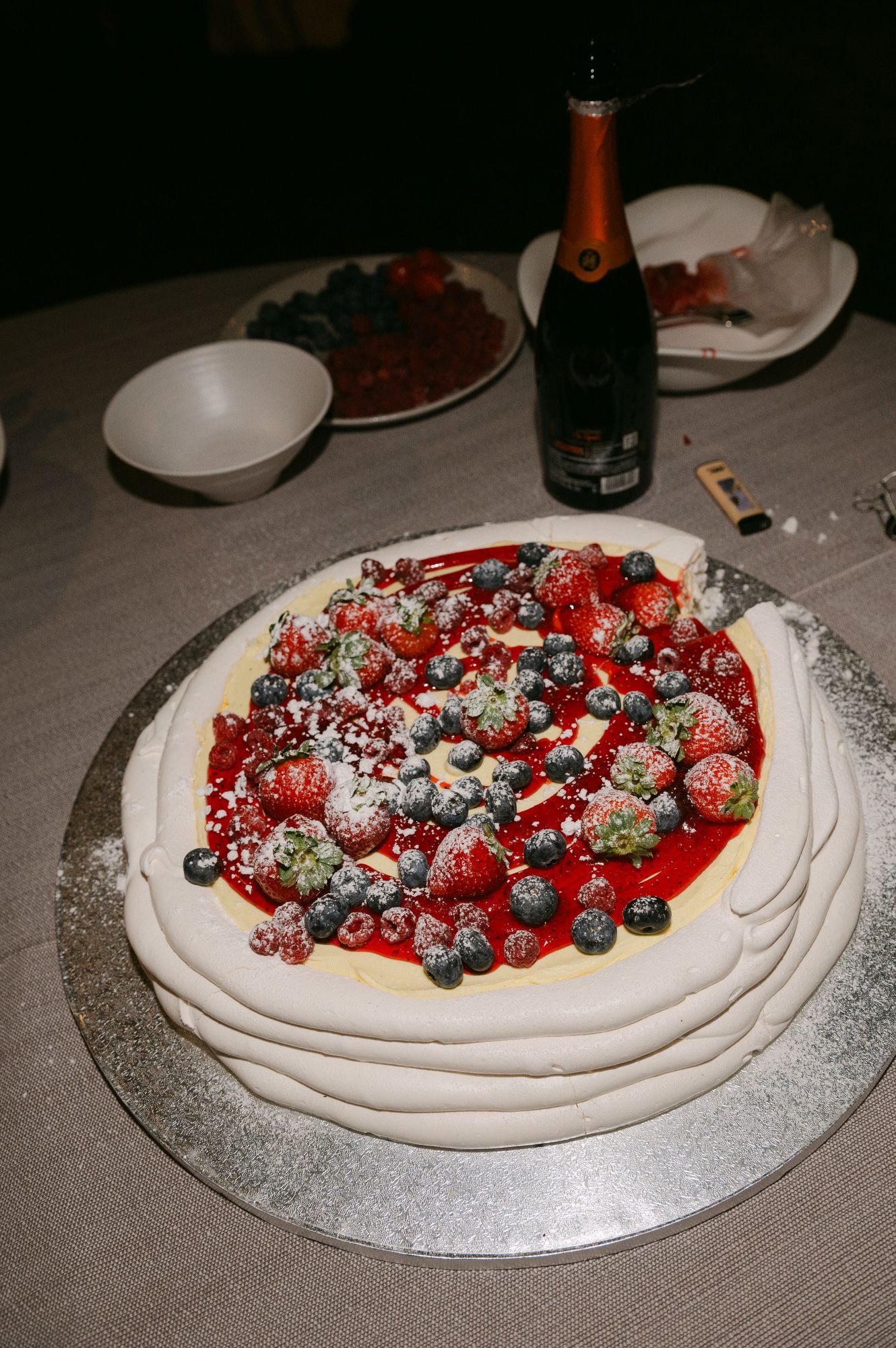 A pavlova dessert topped with fresh strawberries, blueberries and powdered sugar sits on a table with a champagne bottle.