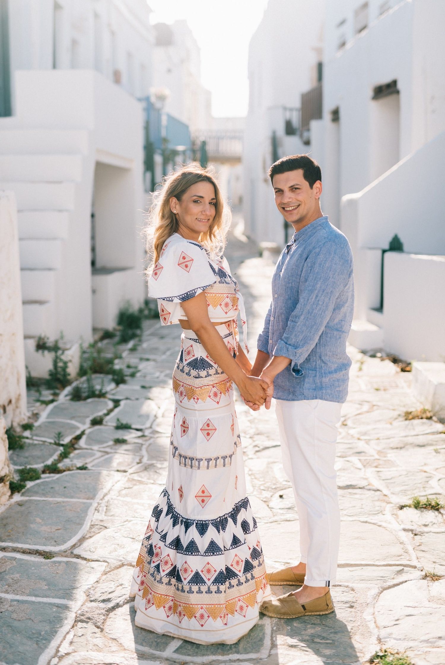 A couple poses in stylish outfits against white Mediterranean architecture.