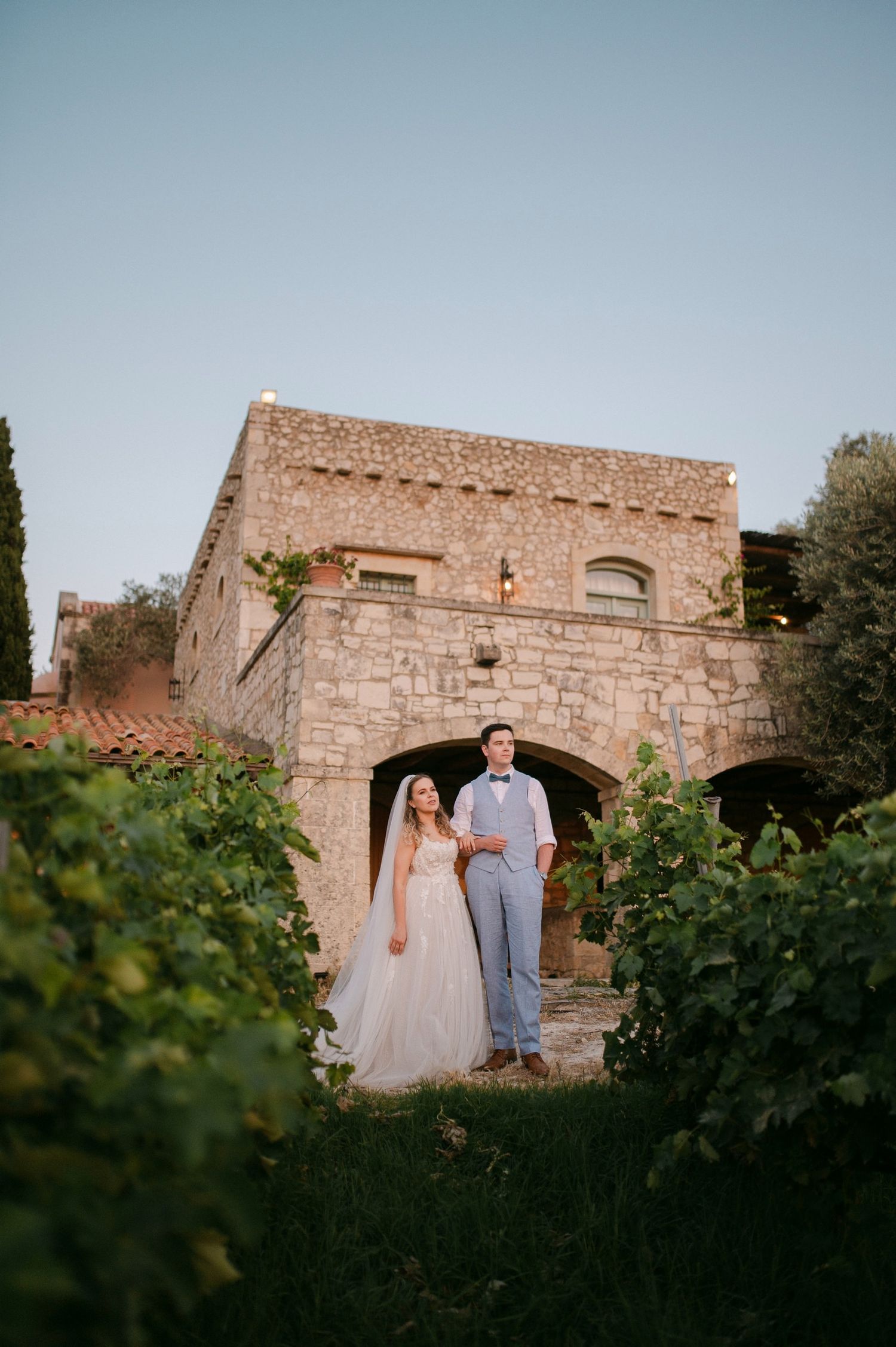 Couple shares romantic moment between vineyard rows at sunset beside a rustic stone building.