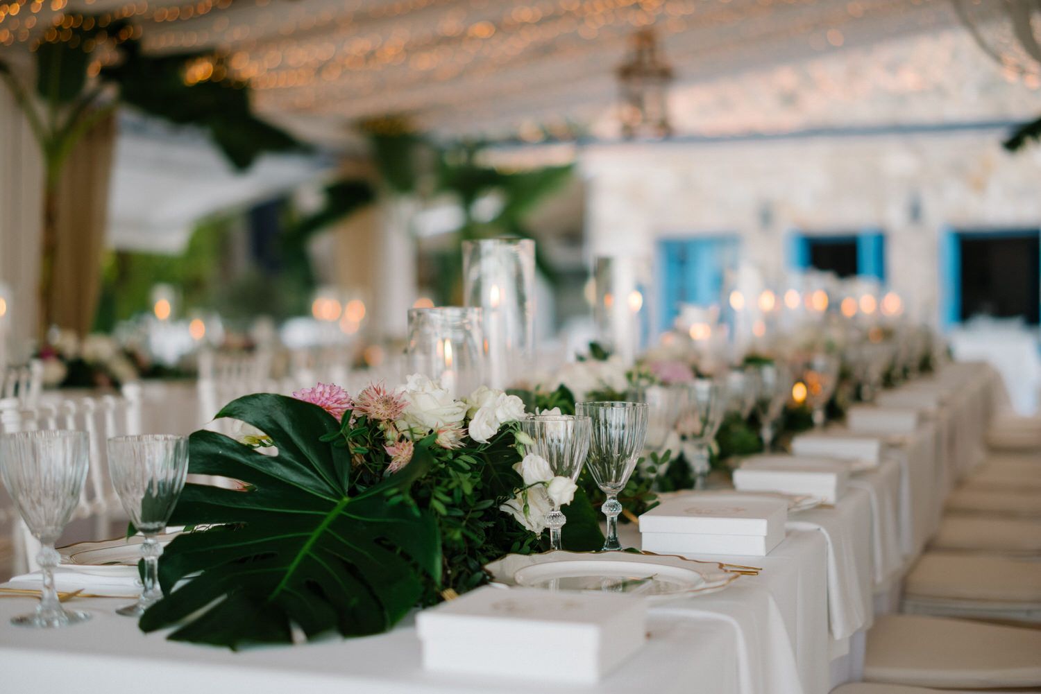 Long wedding reception table decorated with white linens, tropical leaves, and elegant floral centerpieces.