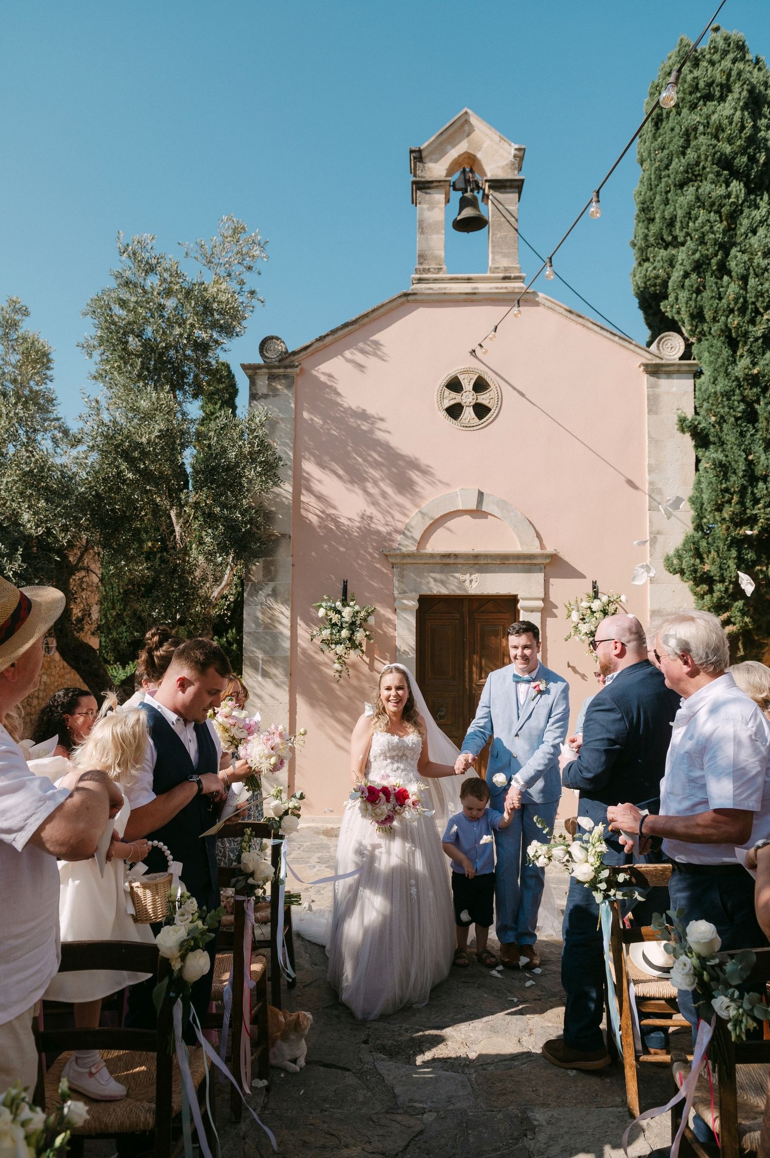 A wedding ceremony takes place outside a charming pink Mediterranean chapel with a bell tower and cypress trees.
