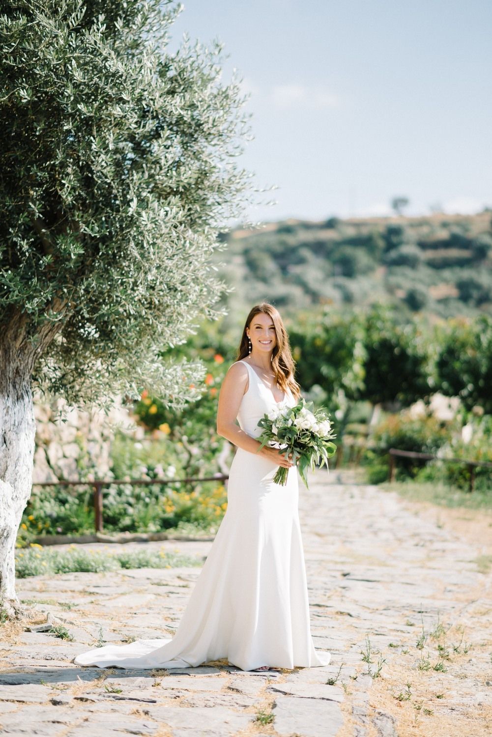 Full-length portrait of bride in sleek white gown holding bouquet in Mediterranean garden setting.