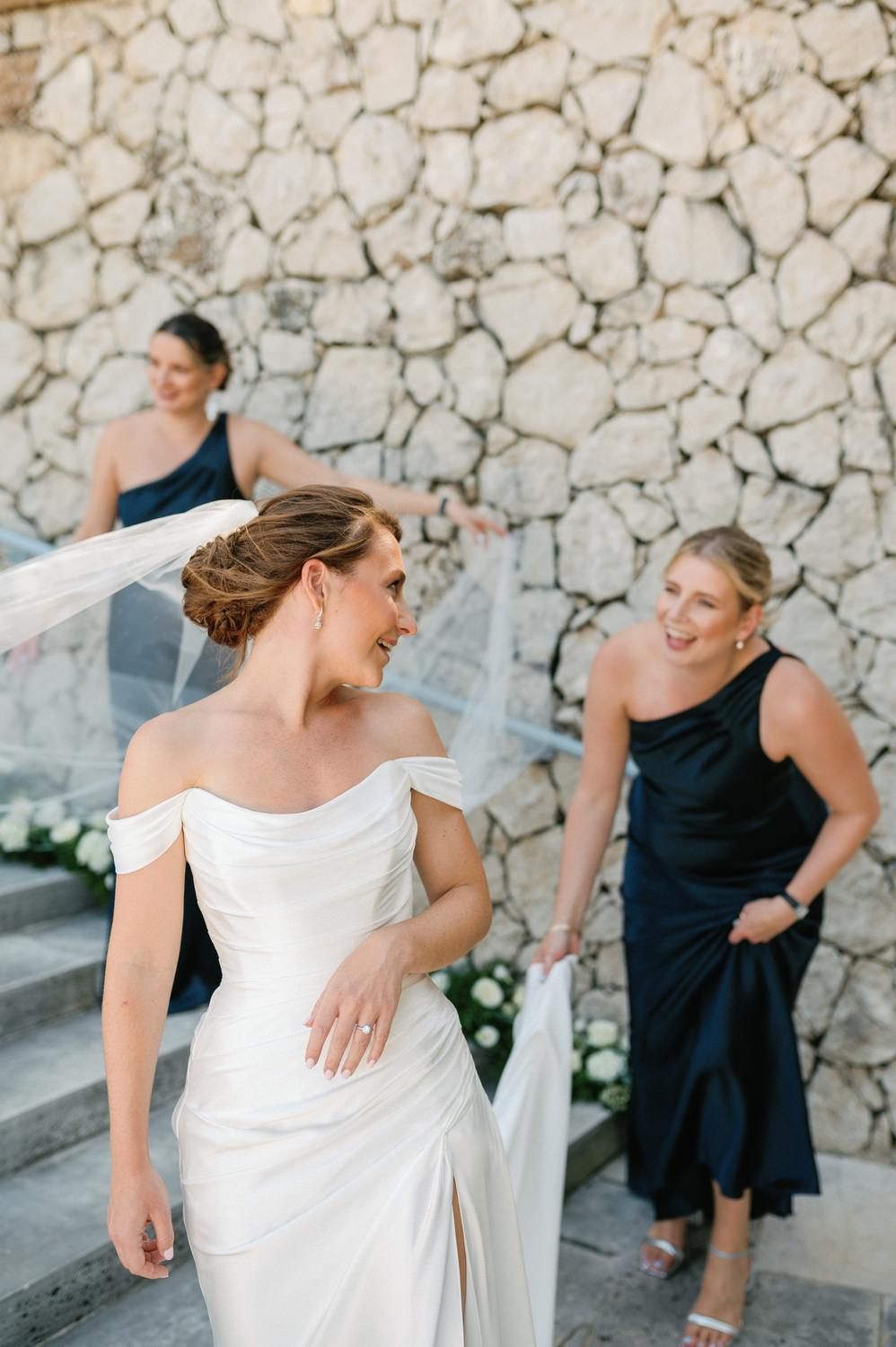 A bride in white and bridesmaids in navy dresses gather on stone steps against a textured wall.