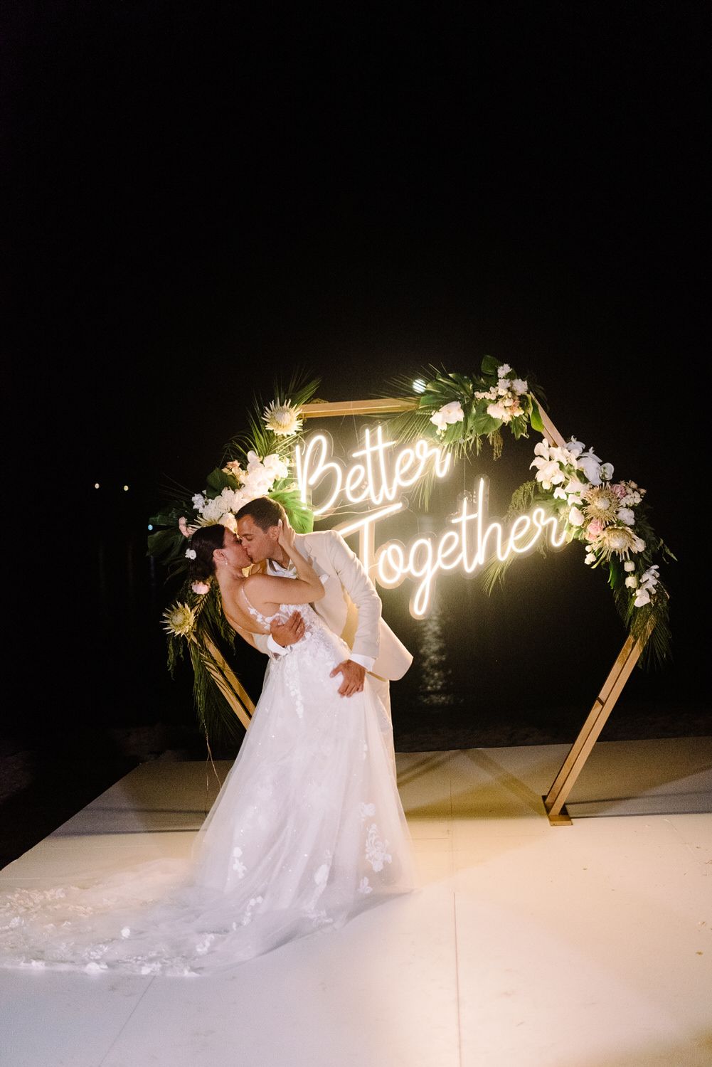 A couple embraces under a glowing hexagonal wedding arch with 'Better Together' neon text and floral decorations.