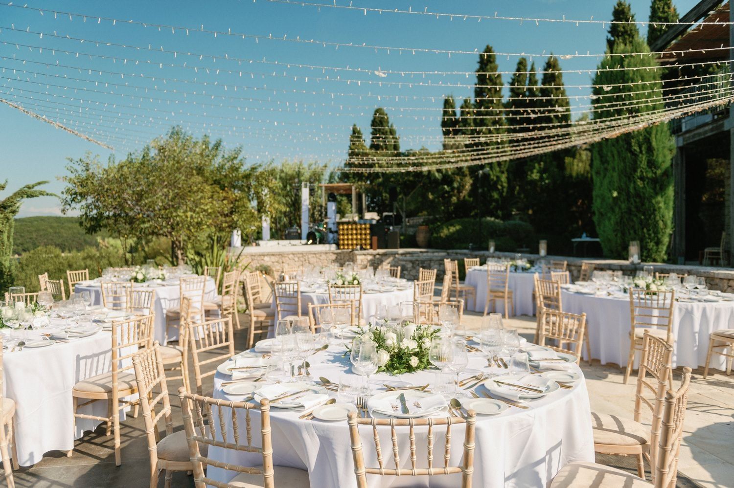 Outdoor wedding reception setup with white tables and string lights under Mediterranean cypress trees.