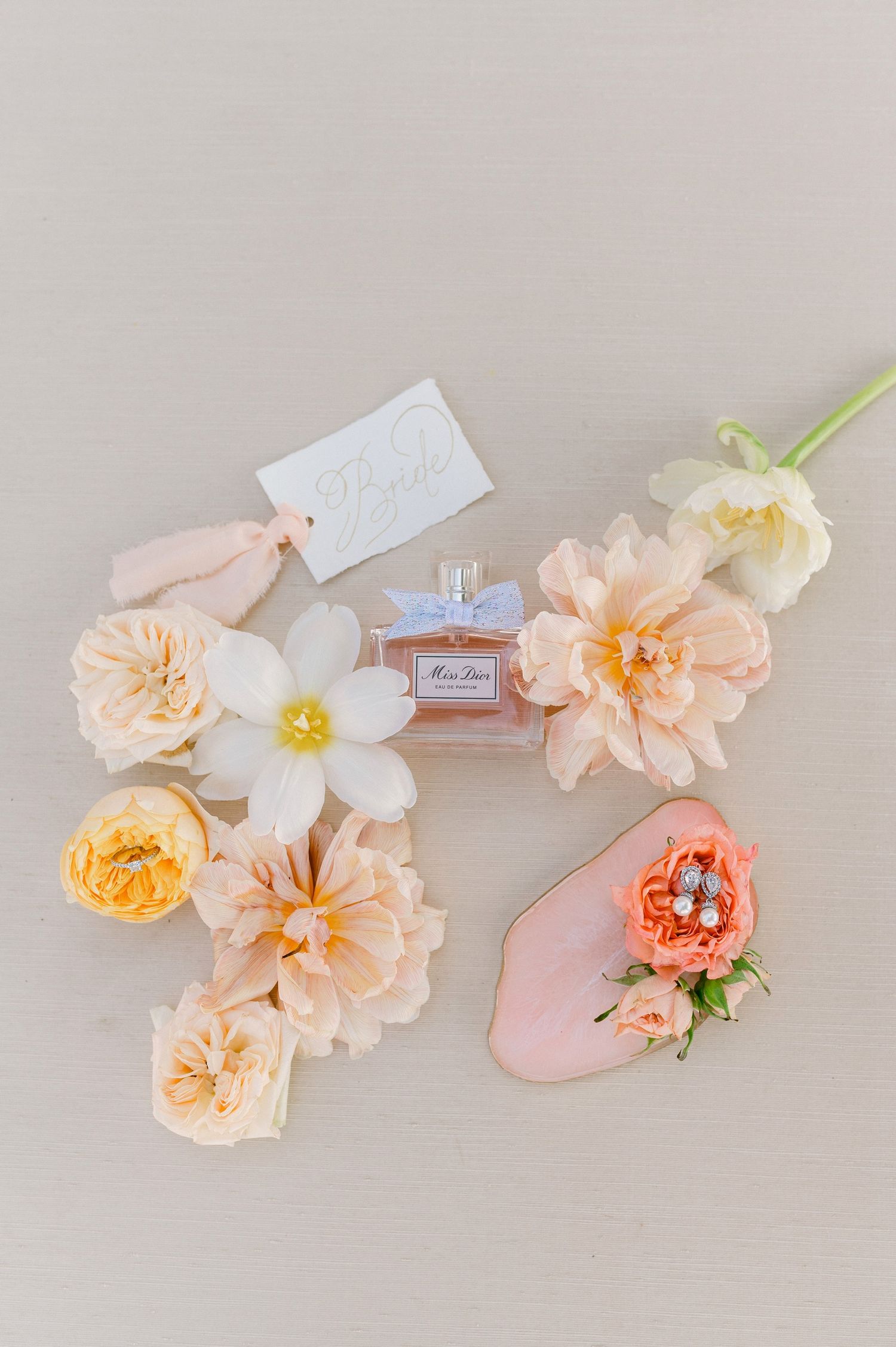 A perfume bottle nestled among delicate peach and cream colored flowers on a light neutral background.