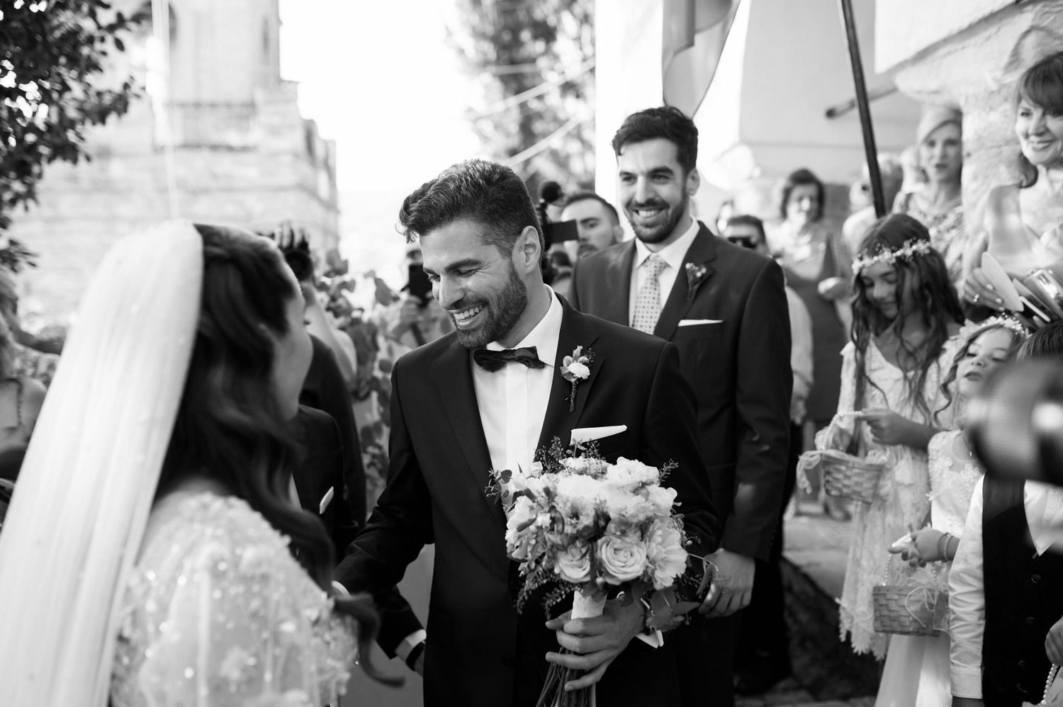 Black and white photo of a joyful wedding moment with newlyweds holding a bouquet surrounded by smiling guests.