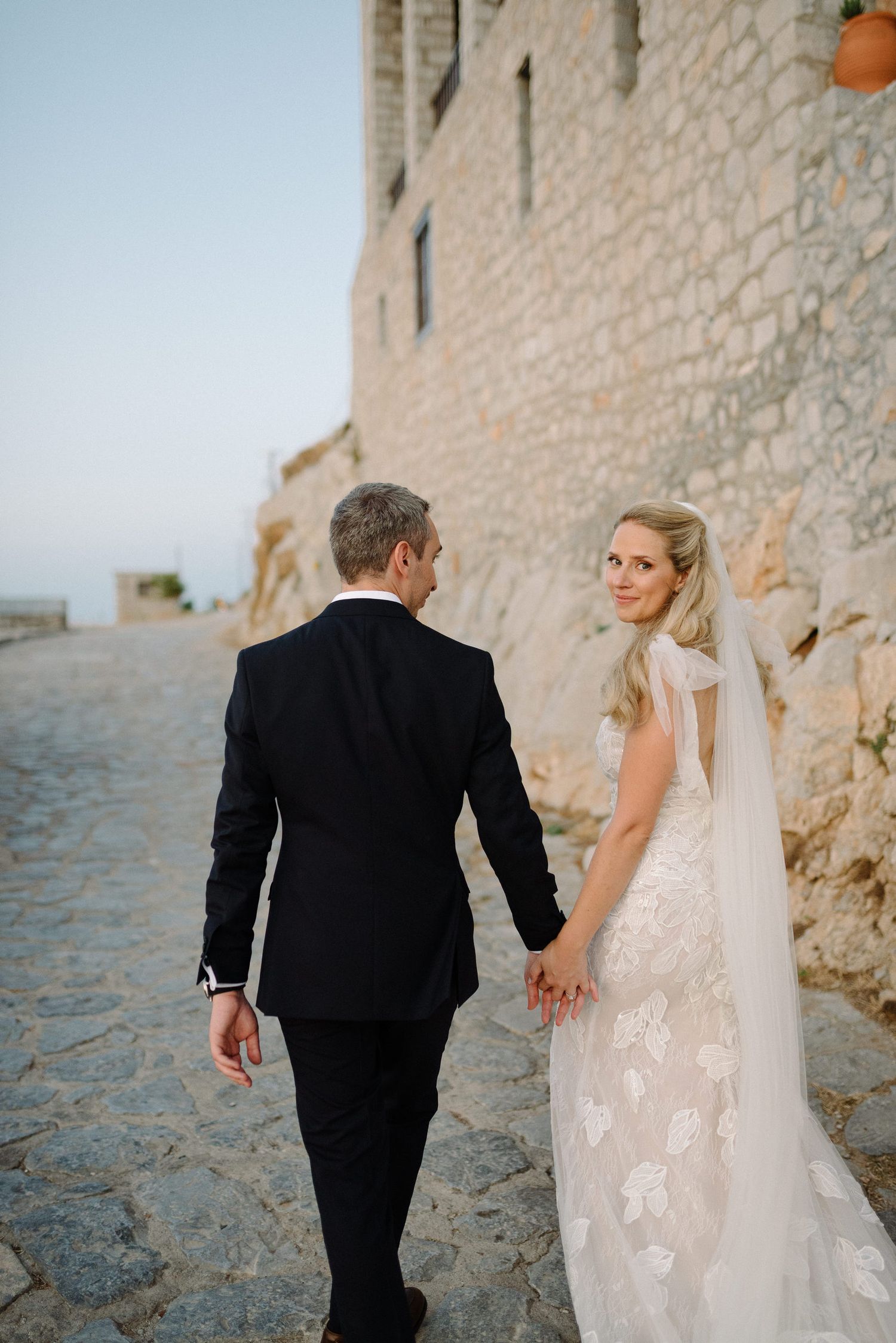A couple in wedding attire walking hand in hand along a stone path in Hydra Greece.