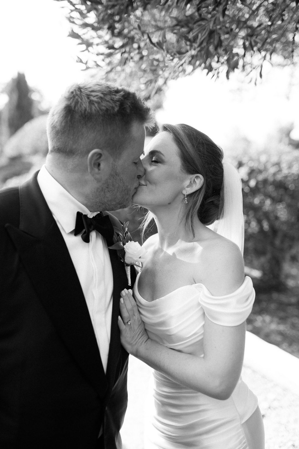 A romantic black and white wedding photo showing a tender kiss between newlyweds under tree branches.