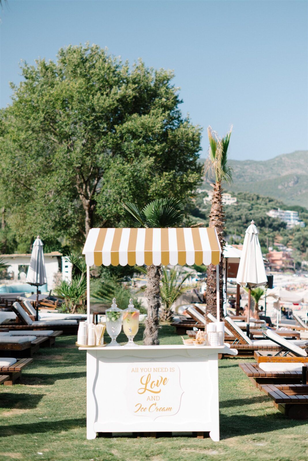 Outdoor beach wedding setup with white cabanas and lounge chairs under striped umbrellas by the ocean.