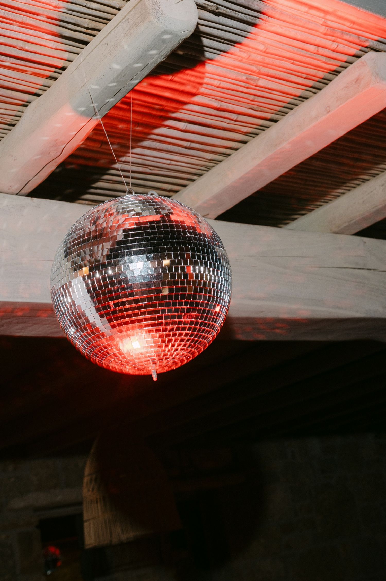 Disco ball hanging from wooden ceiling beams reflects red light in a dark room creating a moody party atmosphere.