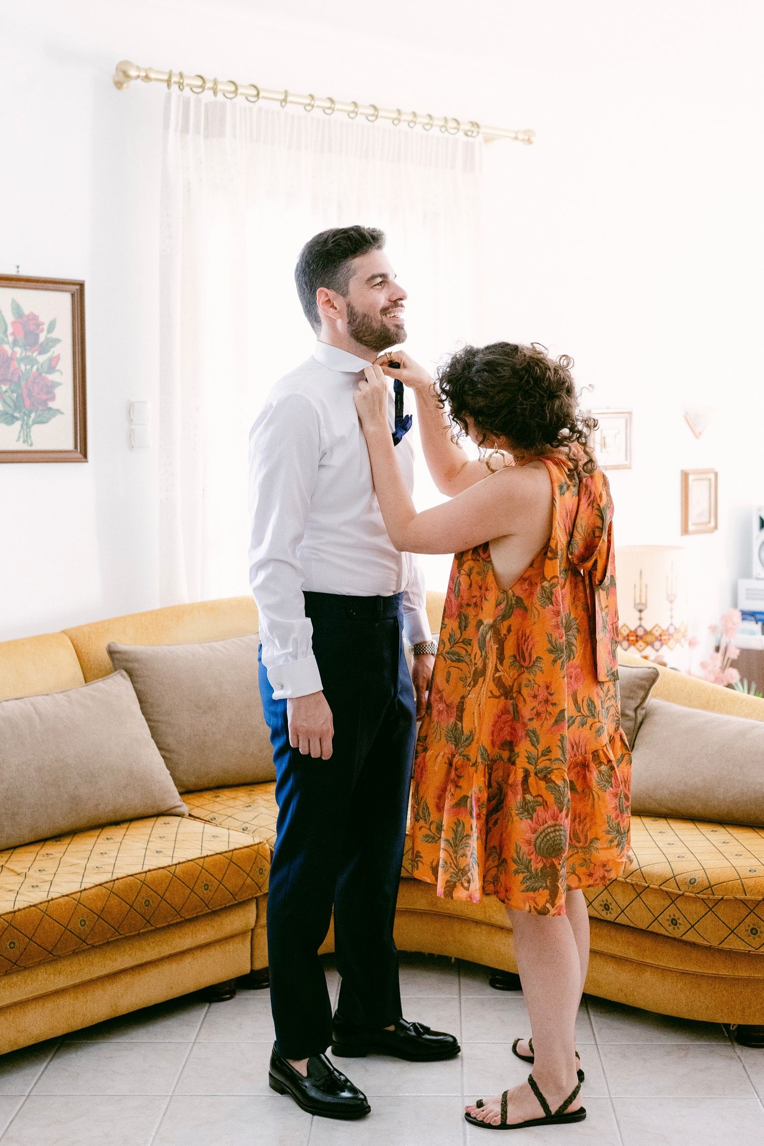 A couple getting ready in a bright living room with a yellow sofa, curtains and framed artwork on the wall.