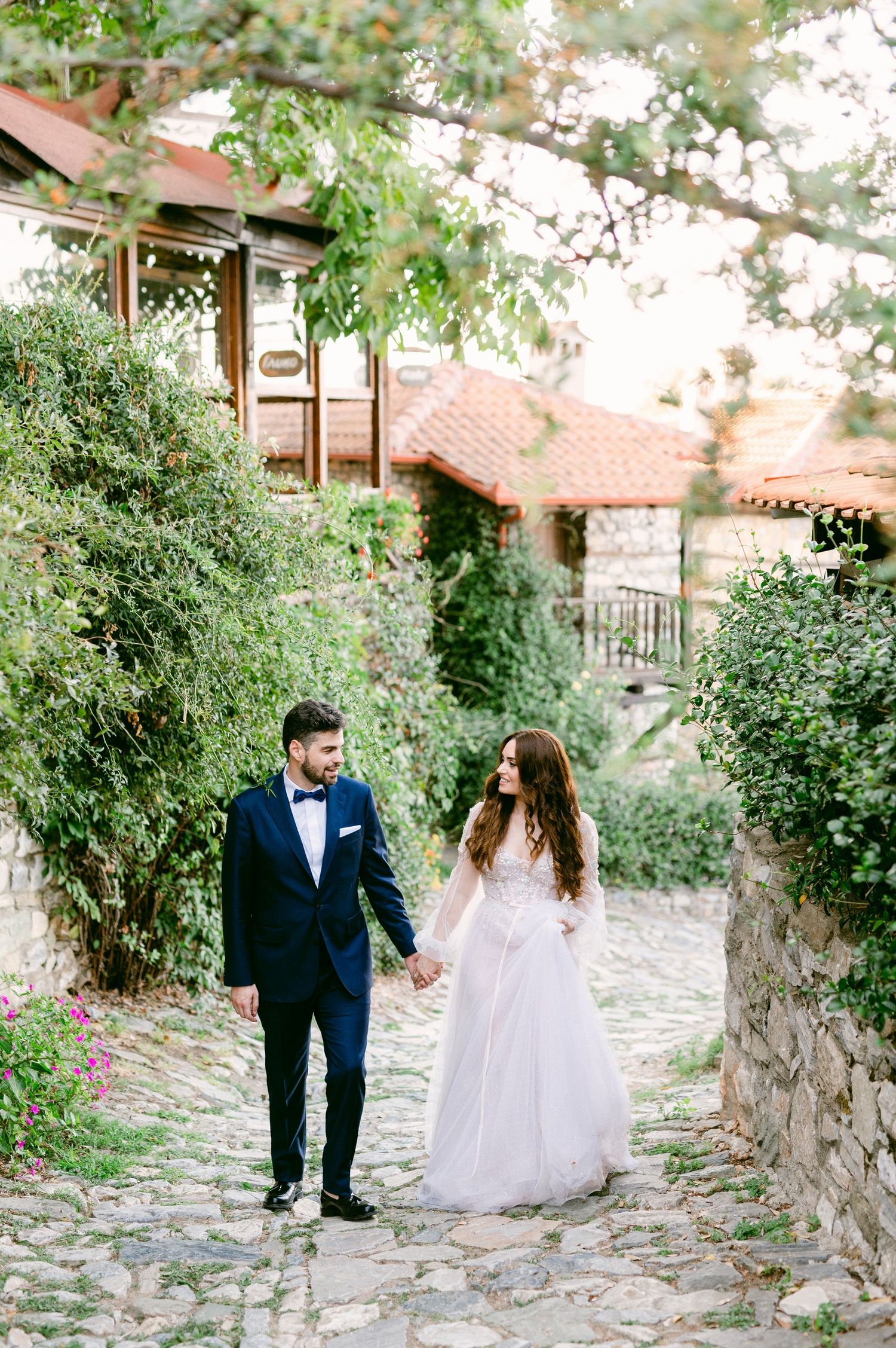 A couple in wedding attire walks hand in hand on a charming cobblestone path surrounded by lush greenery and rustic buildings.