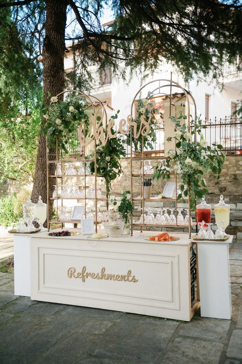 White refreshment station with greenery decorations and drink dispensers at an outdoor wedding.