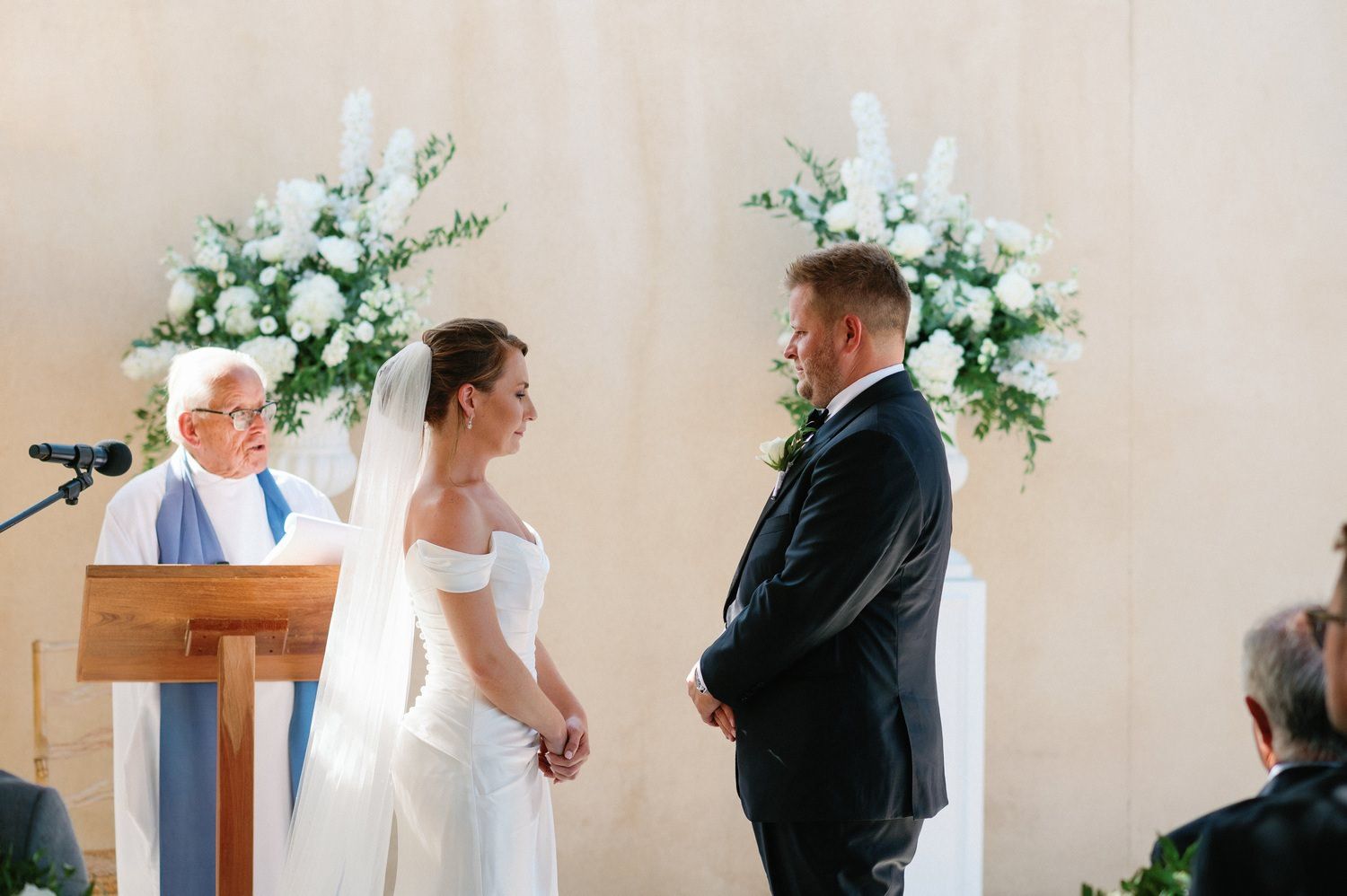 Elegant wedding ceremony with two standing before an officiant with white floral arrangements in the background.