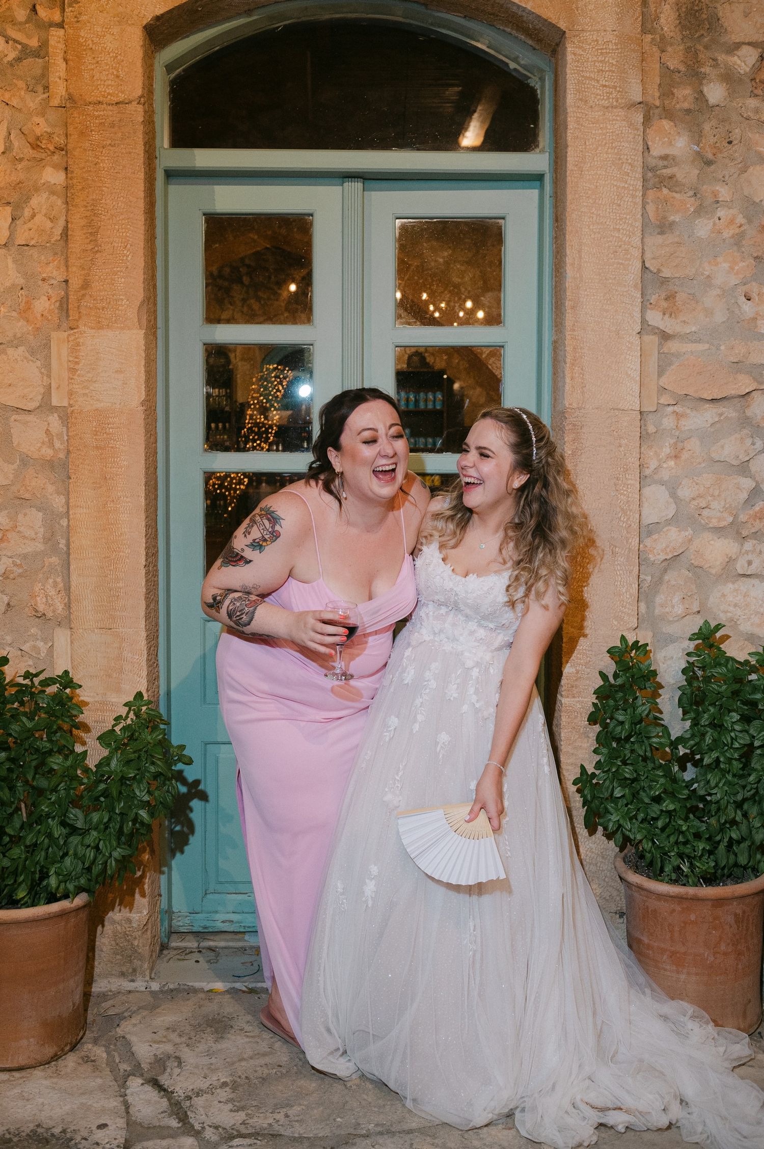 Two people share a joyful laugh together outside a turquoise door at a wedding celebration.