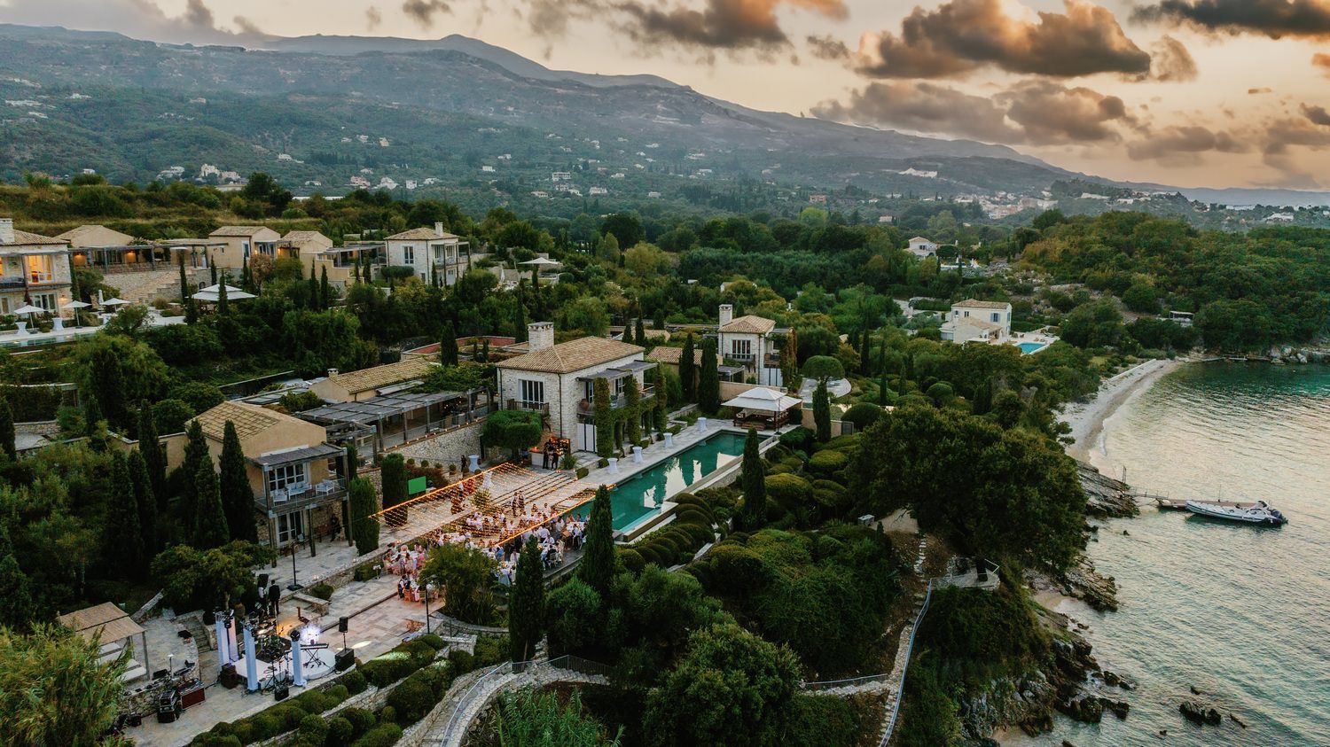 Aerial view of a coastal wedding venue with infinity pool and sunset over the mountains.