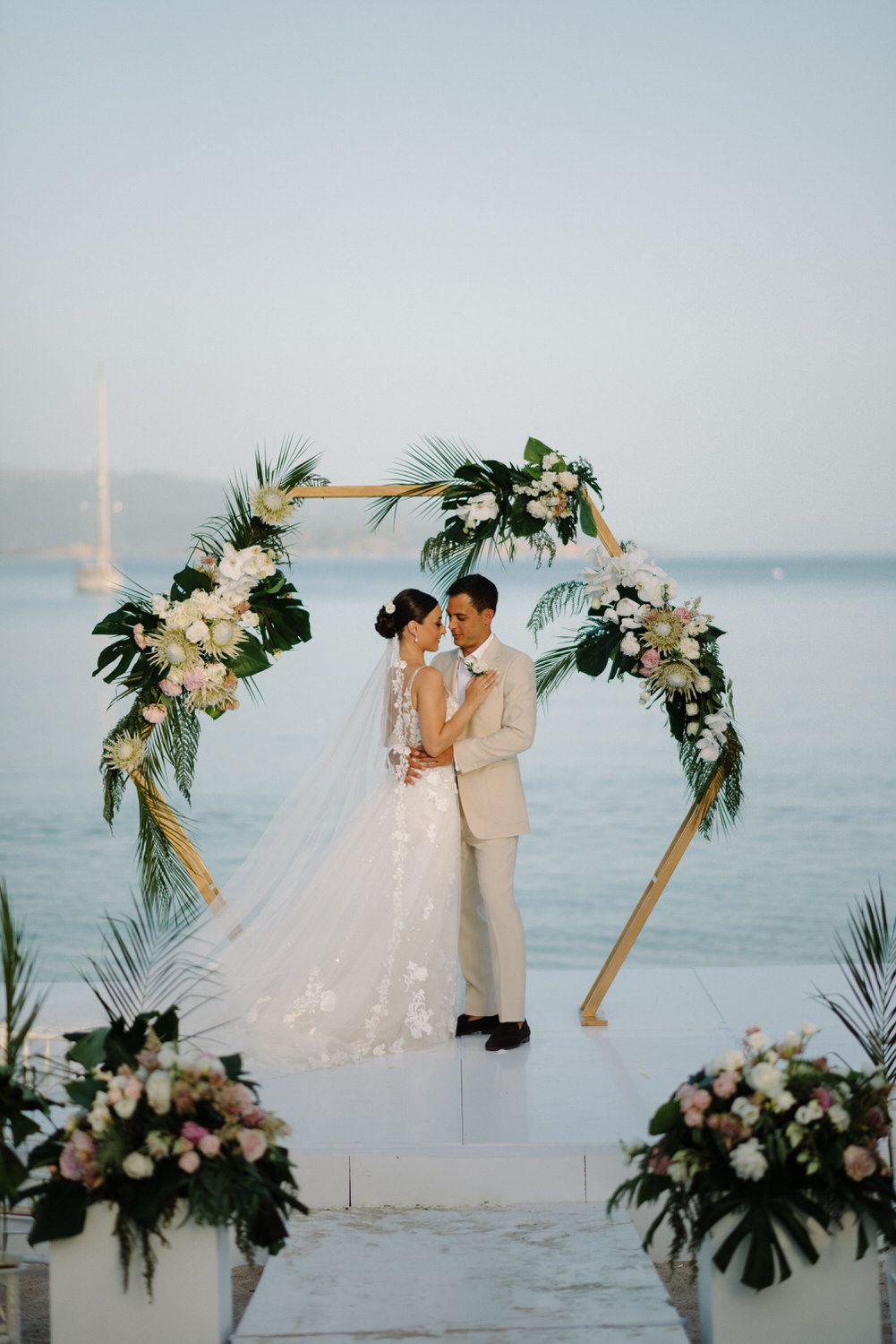 Wedding couple shares romantic moment under floral arch at sunset beach ceremony.