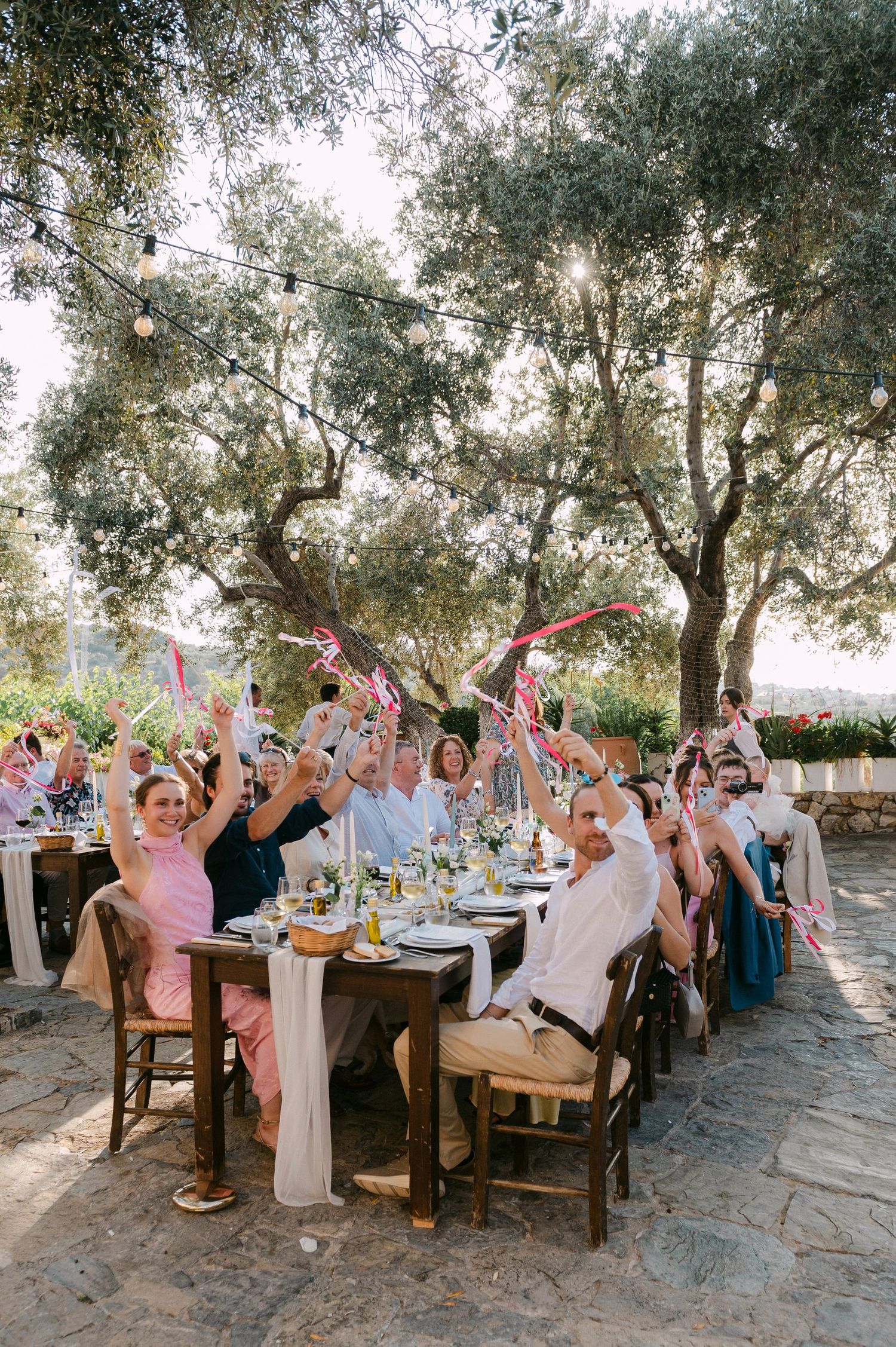 A lively outdoor wedding reception celebration with guests seated at long rustic tables under olive trees.