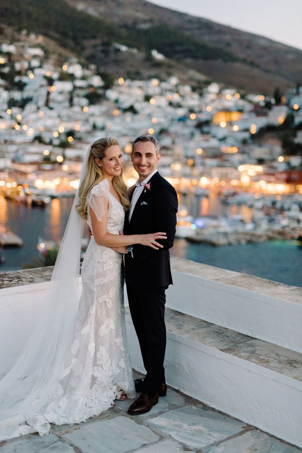 Wedding portrait on white terrace overlooking seaside town with boats in harbor.