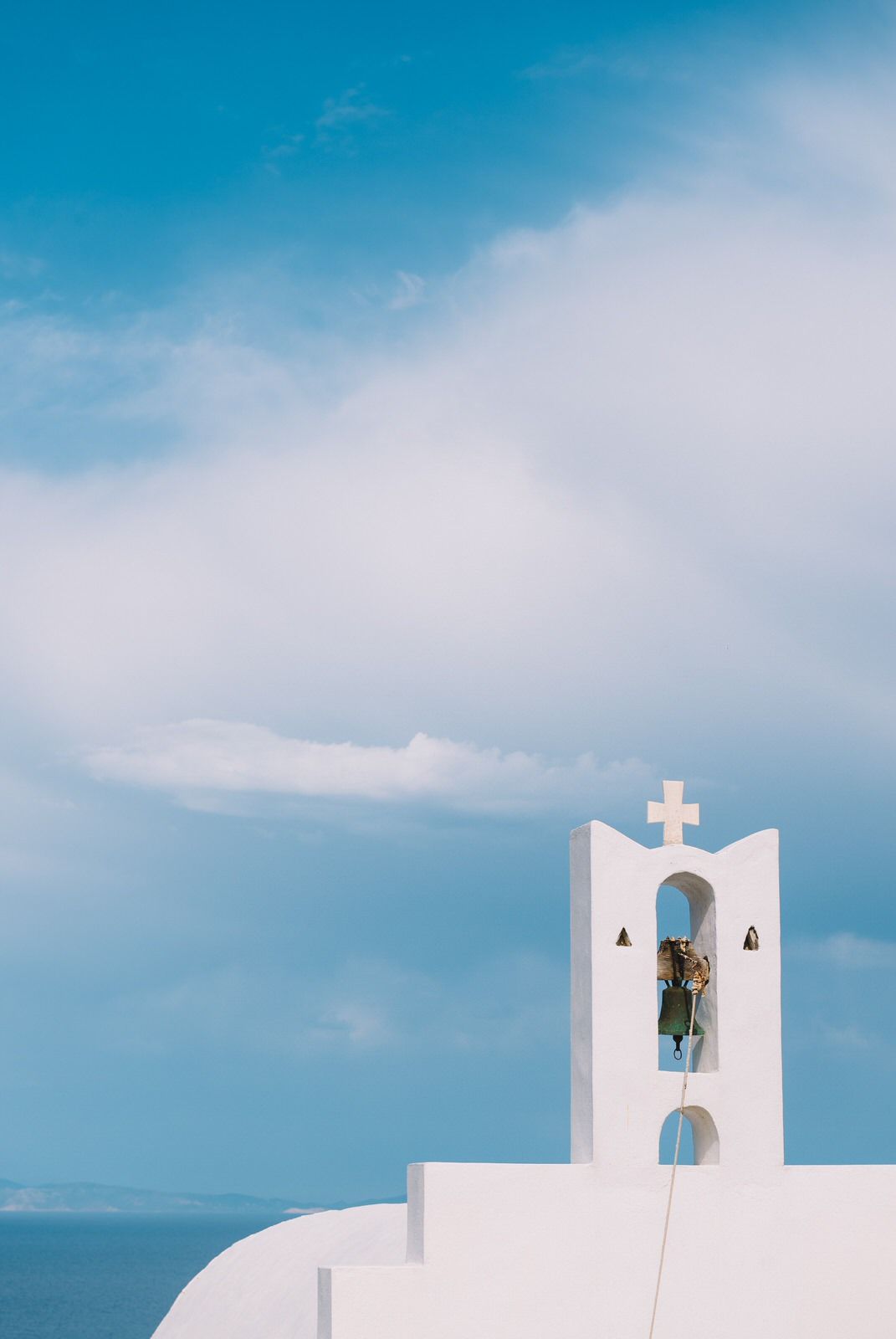 A white Greek Orthodox church bell tower with cross against bright blue sky and ocean in Santorini, Greece.