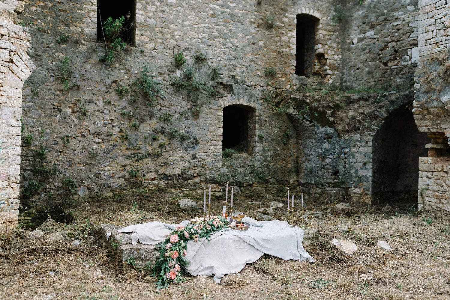 Elegant picnic setup against rustic stone castle wall with white linens and floral decorations.