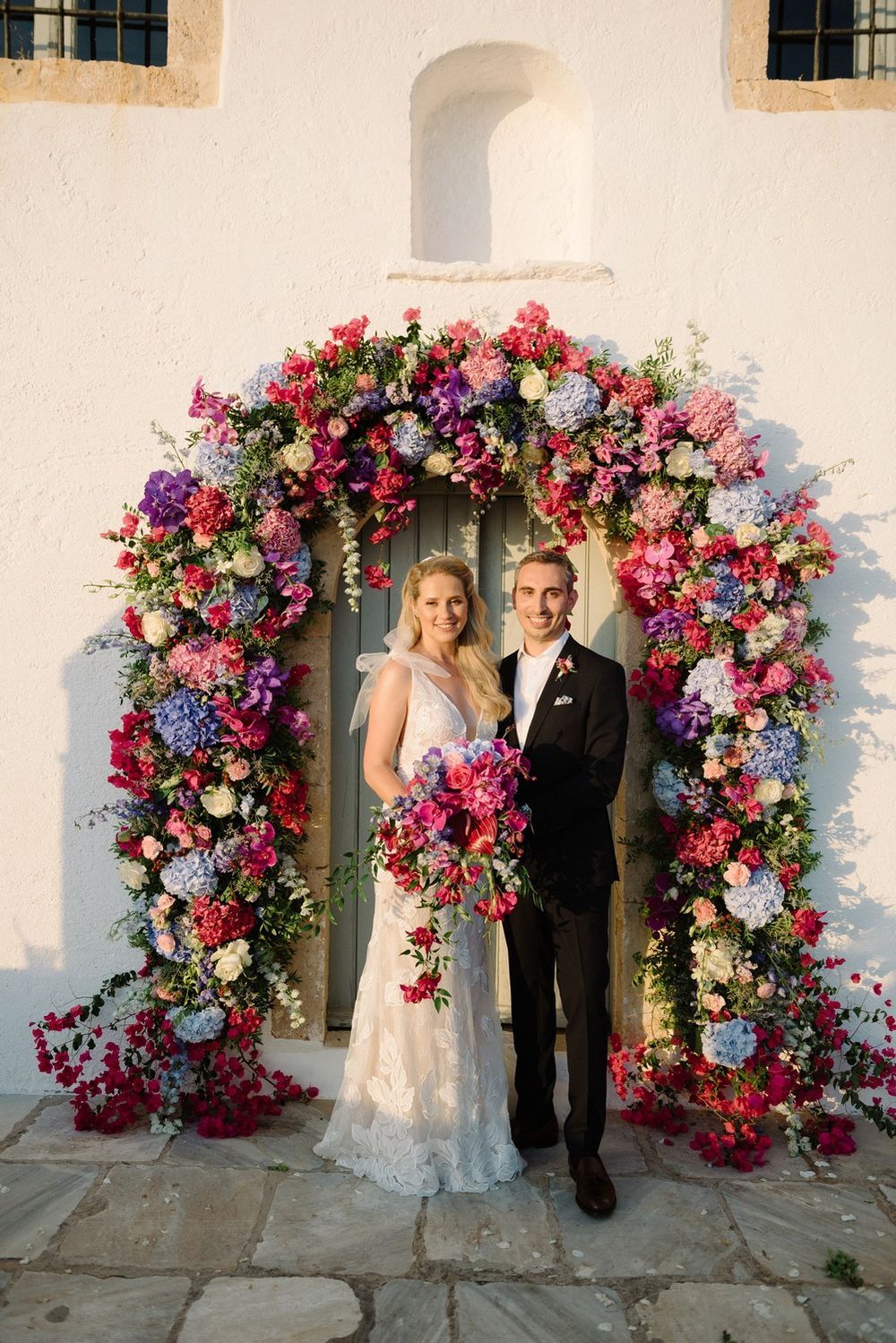 A stunning floral arch with pink and blue blooms frames a romantic wedding scene in Hydra, Greece.