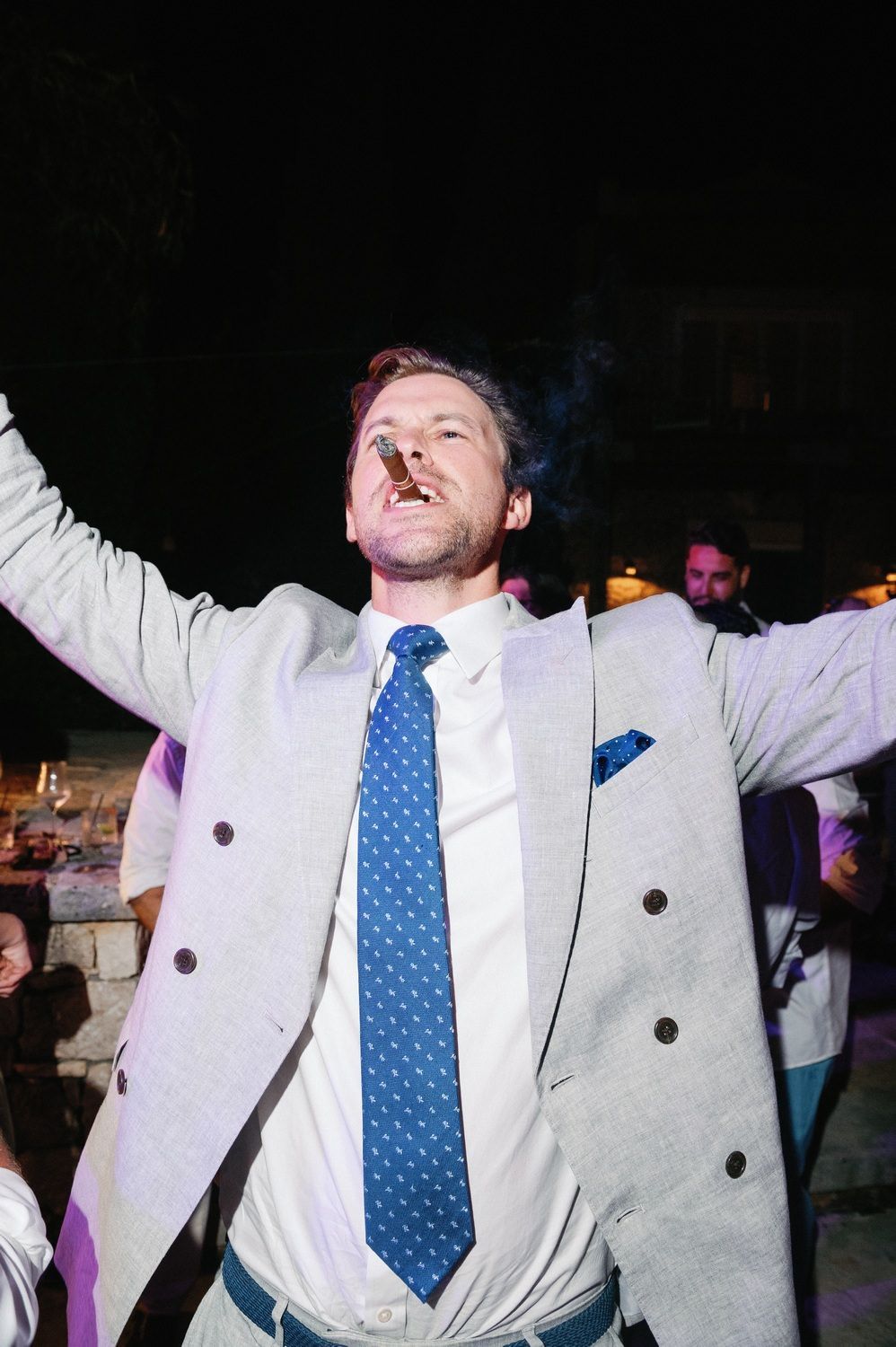 A person in a white double-breasted suit and blue polka dot tie dances energetically with arms raised at a party.