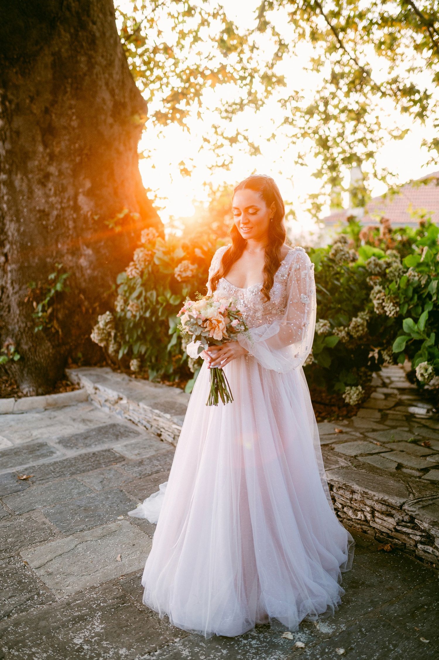 A bride in a long-sleeved lace wedding gown holds a bouquet at sunset with golden light filtering through trees.