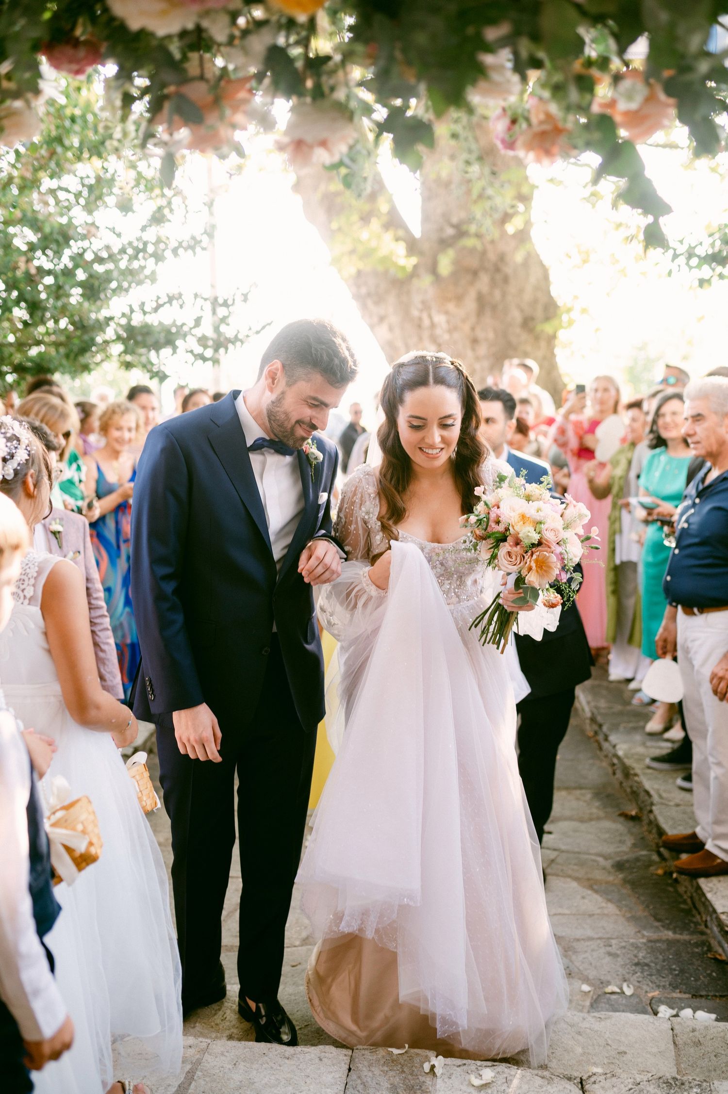 A couple celebrating their wedding day walking together outdoors under floral decorations as guests look on.
