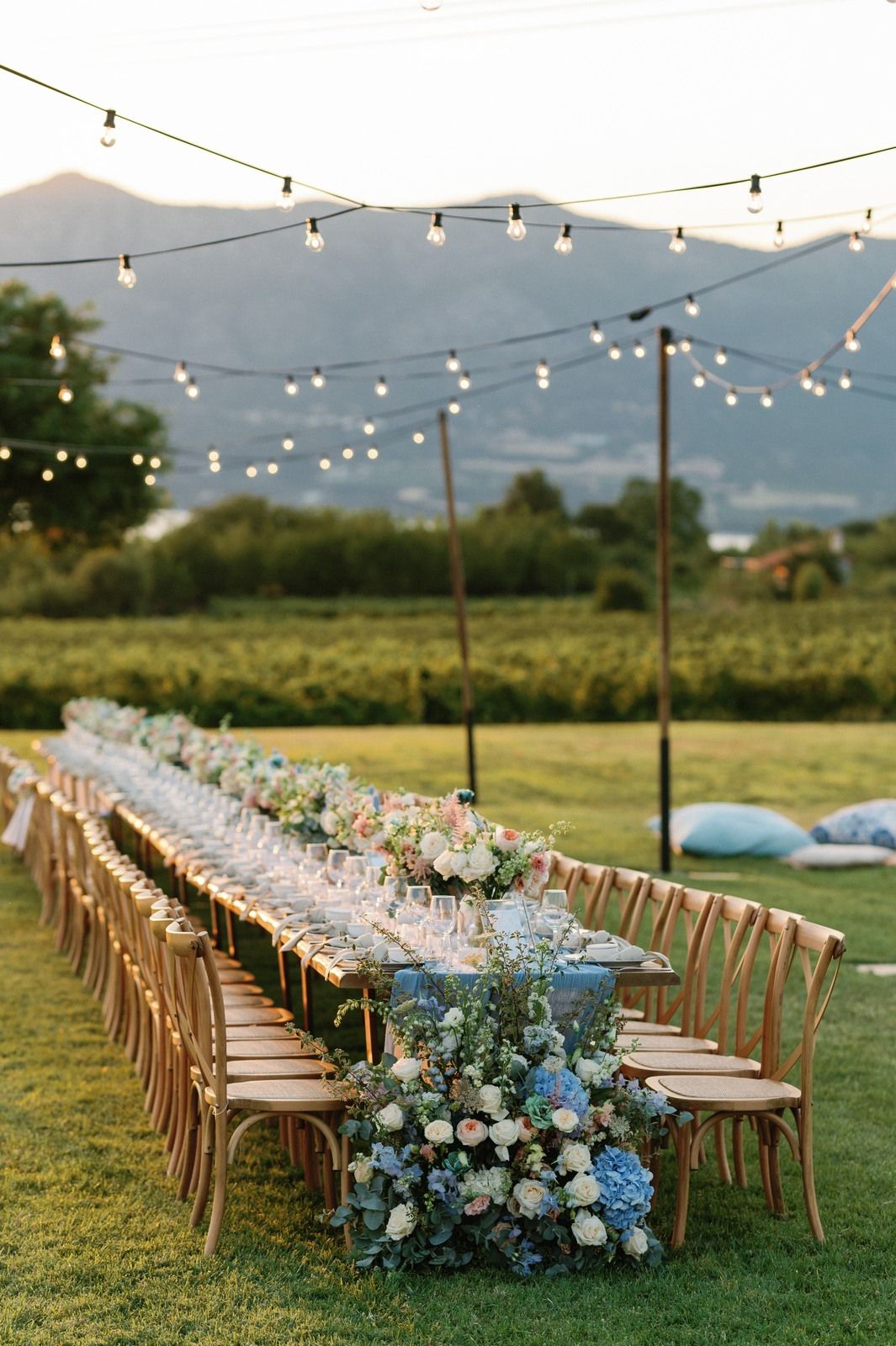 Long banquet table set for outdoor wedding reception with blue floral runner and string lights over mountain landscape.