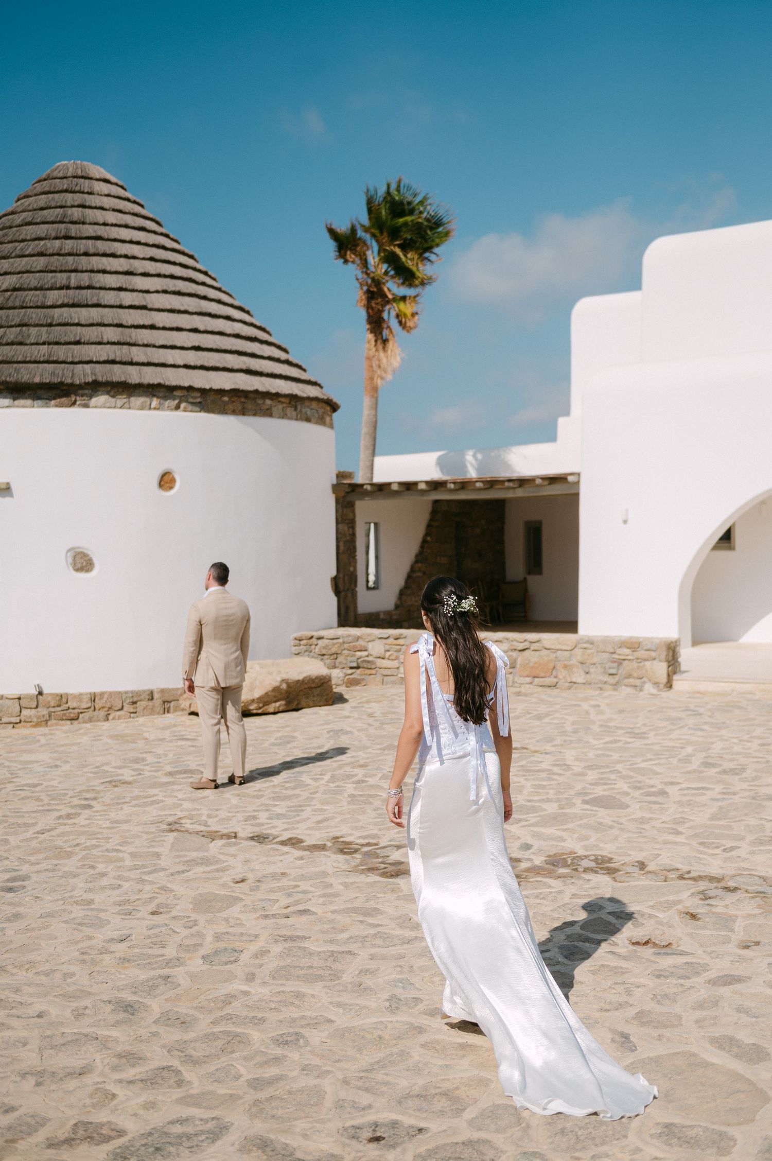 A figure in a flowing white dress walks through a Mediterranean architectural setting with domed buildings and sandy paths.