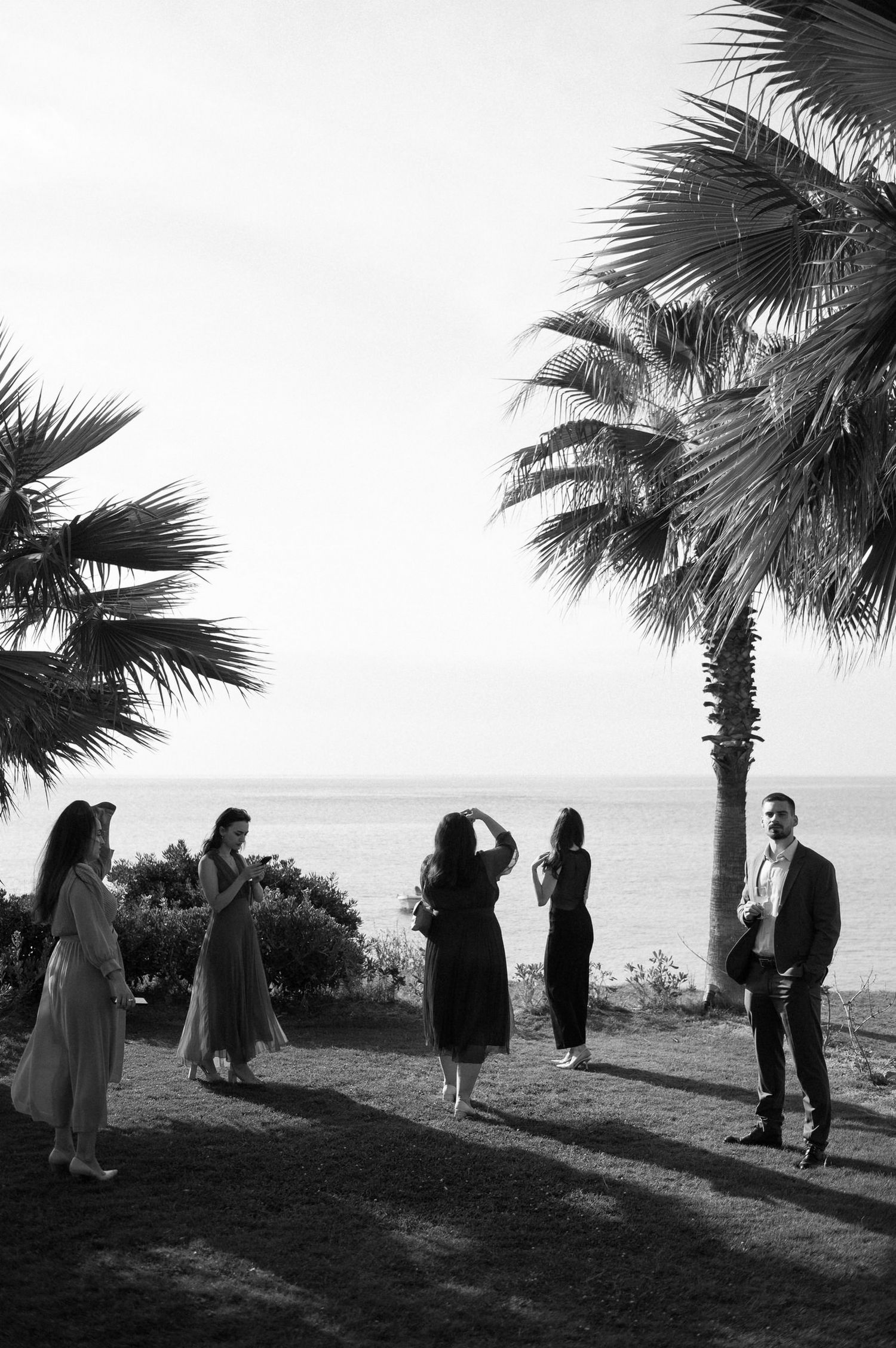A black and white photo of guests gathered under palm trees at a seaside event.