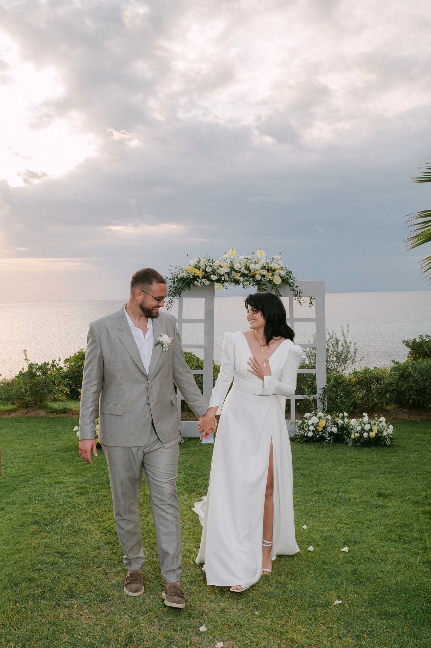 A couple in wedding attire walks together on a grassy lawn by the ocean during an overcast day.