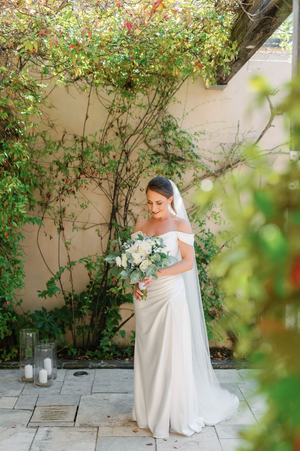 A bride in an off-shoulder gown holds a white bouquet while standing beside climbing vines and garden decor.