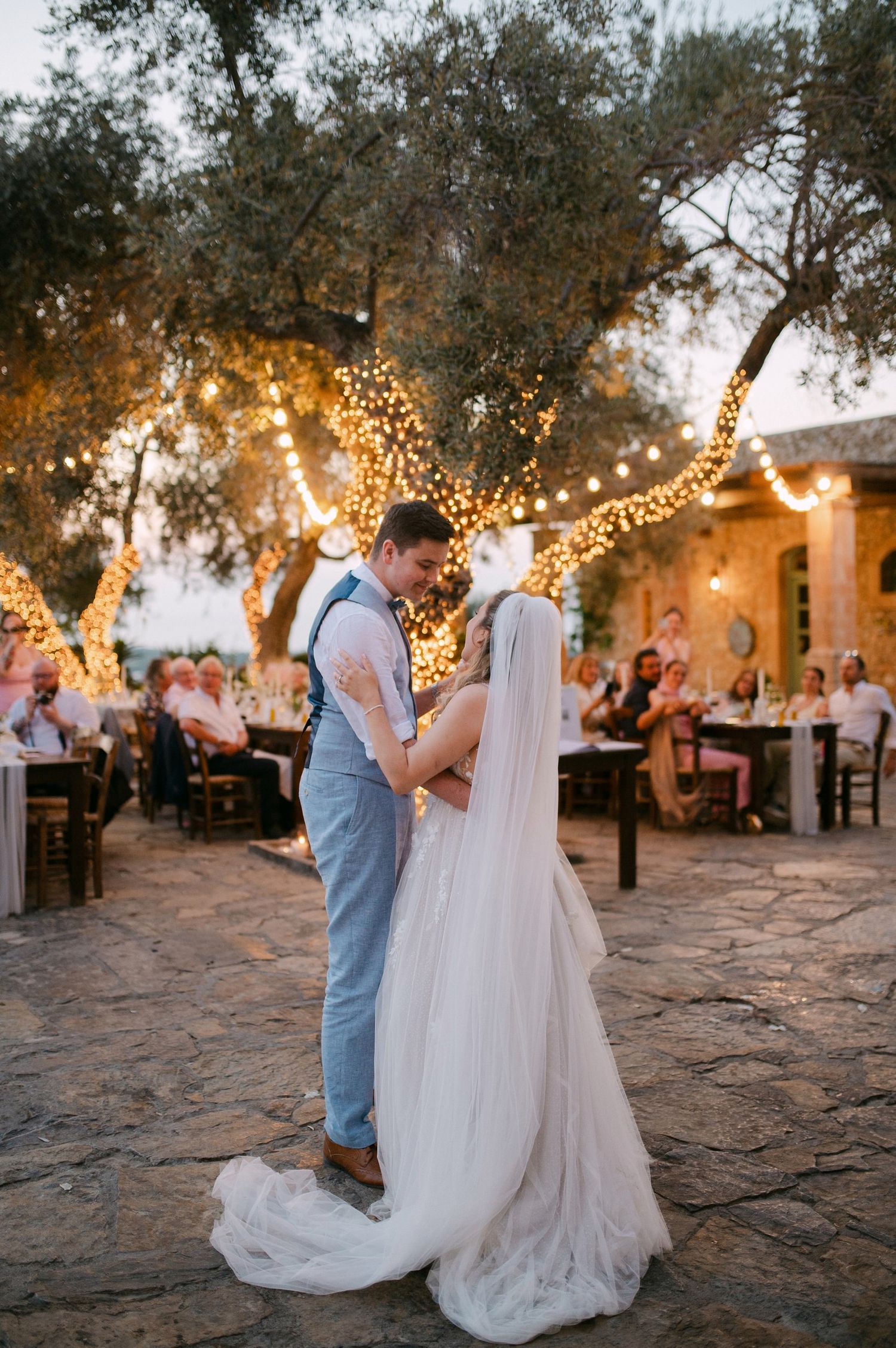 A romantic evening wedding celebration with string lights illuminating olive trees over a stone courtyard.