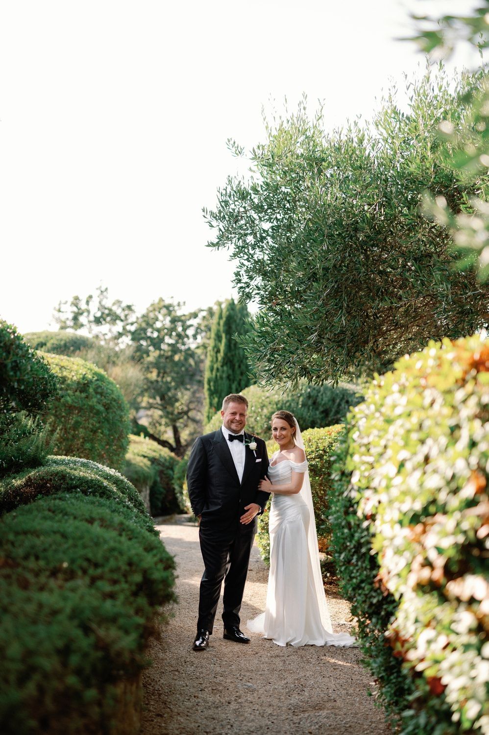 Wedding couple standing on garden path surrounded by olive trees and manicured hedges.