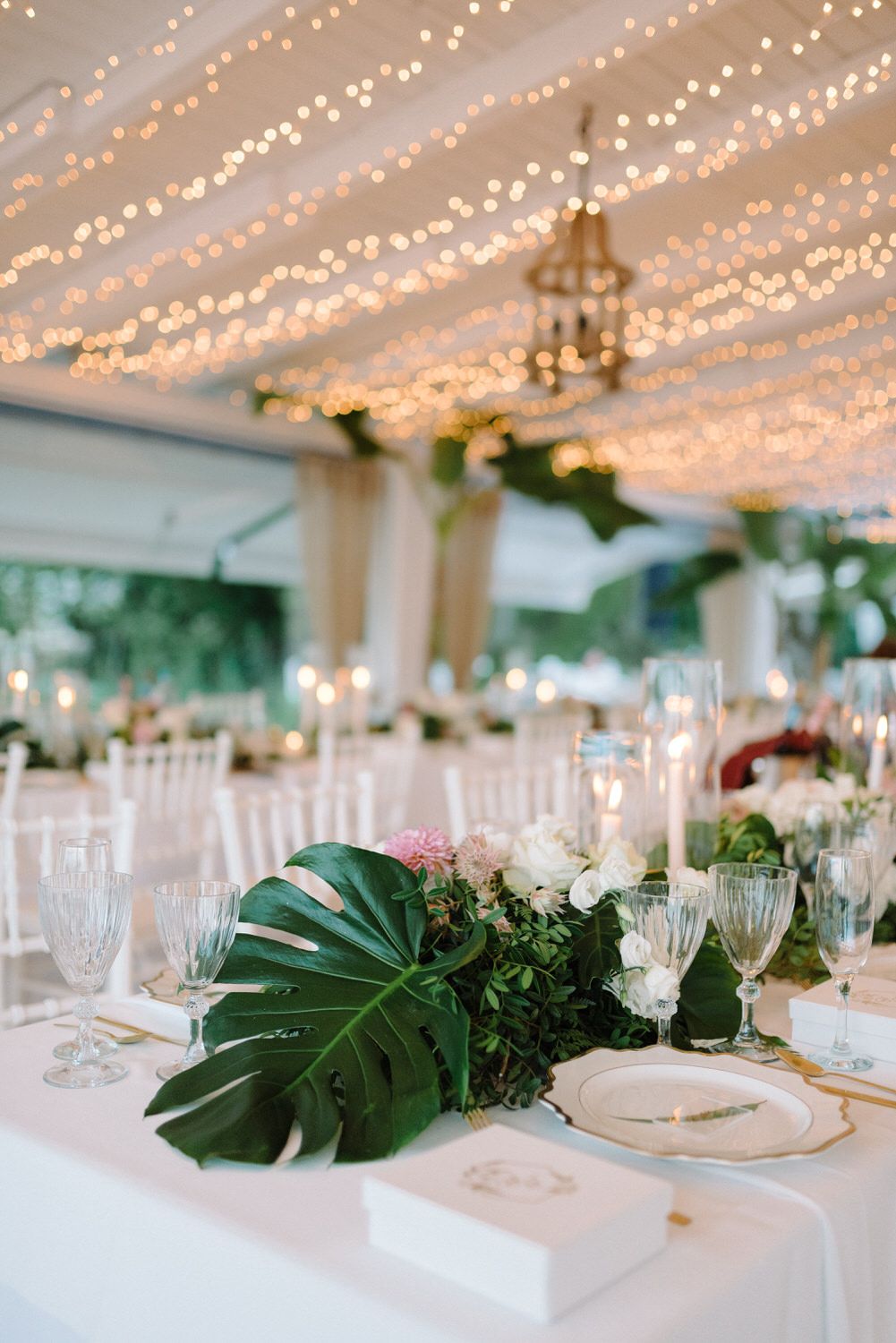 Elegant wedding table setting under twinkling fairy lights with tropical greenery and crystal glassware.
