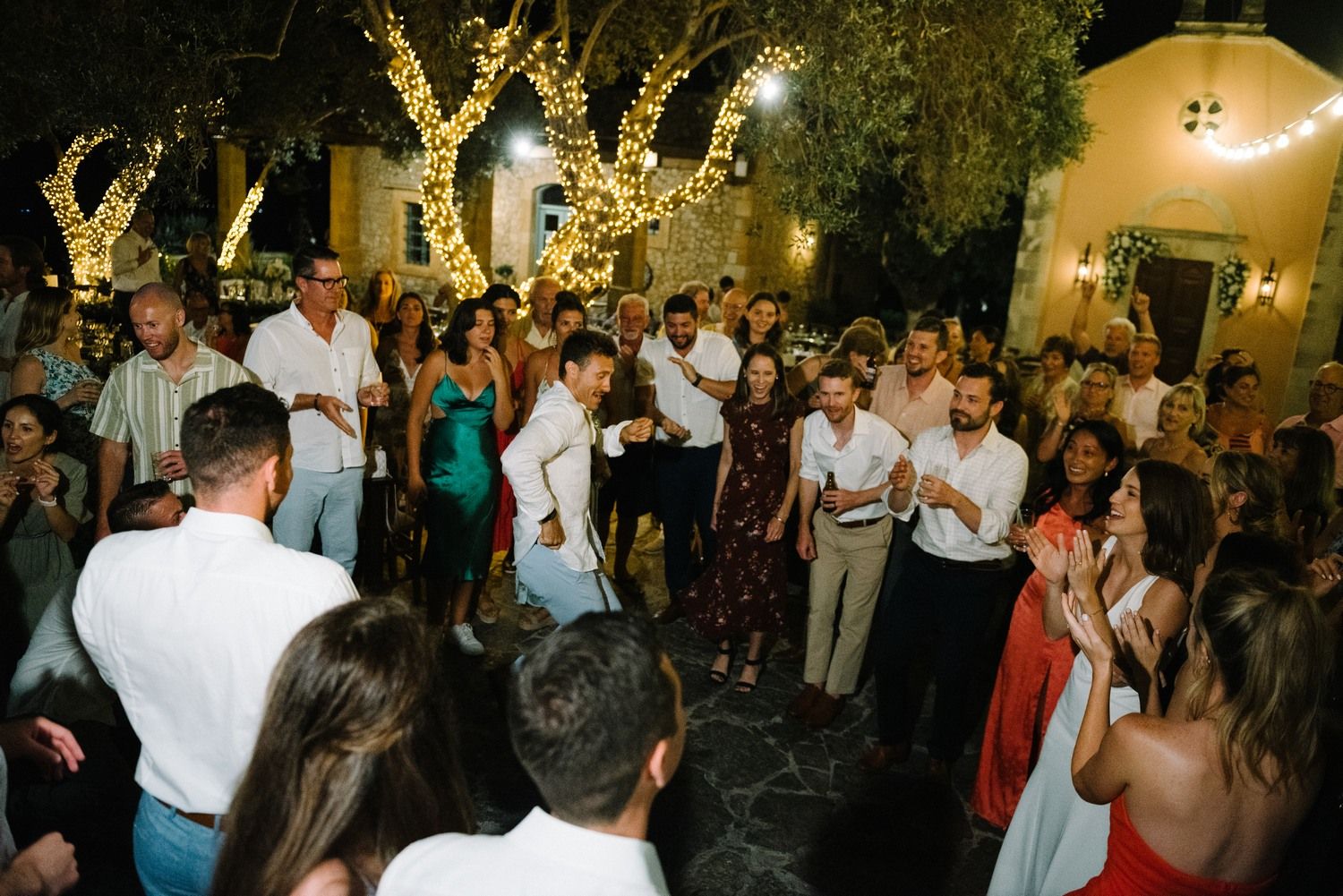 Wedding guests dancing and celebrating at night under illuminated trees.